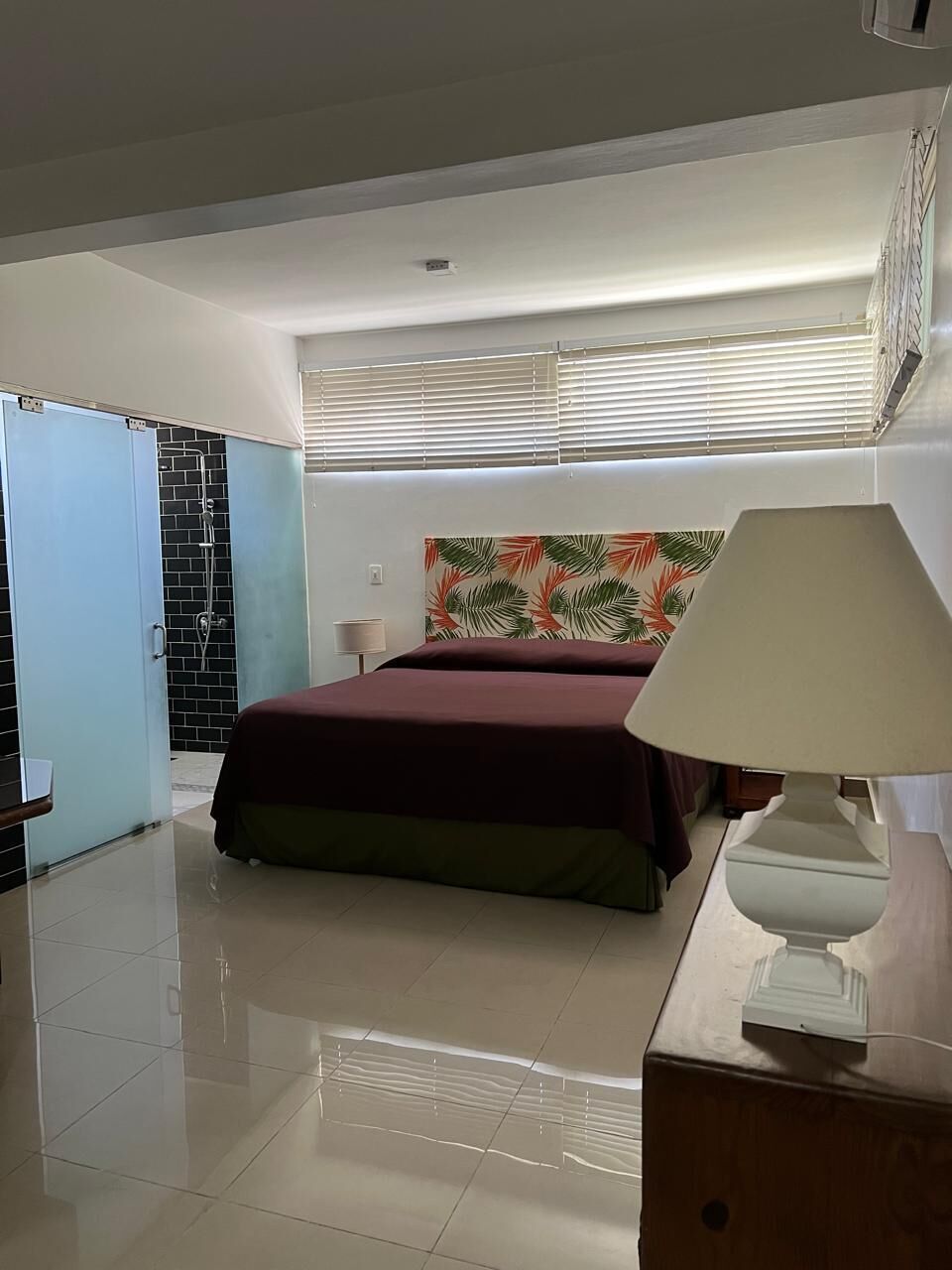 A bedroom with a bed, a nightstand, a lamp, a shower area, and a window with blinds.