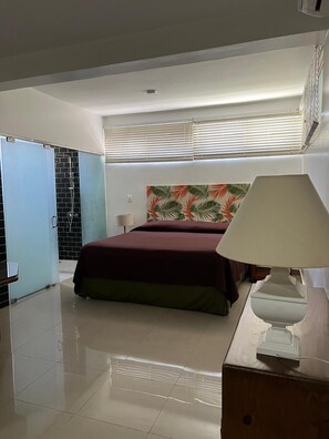 A bedroom with a bed, a nightstand, a lamp, a shower area, and a window with blinds.