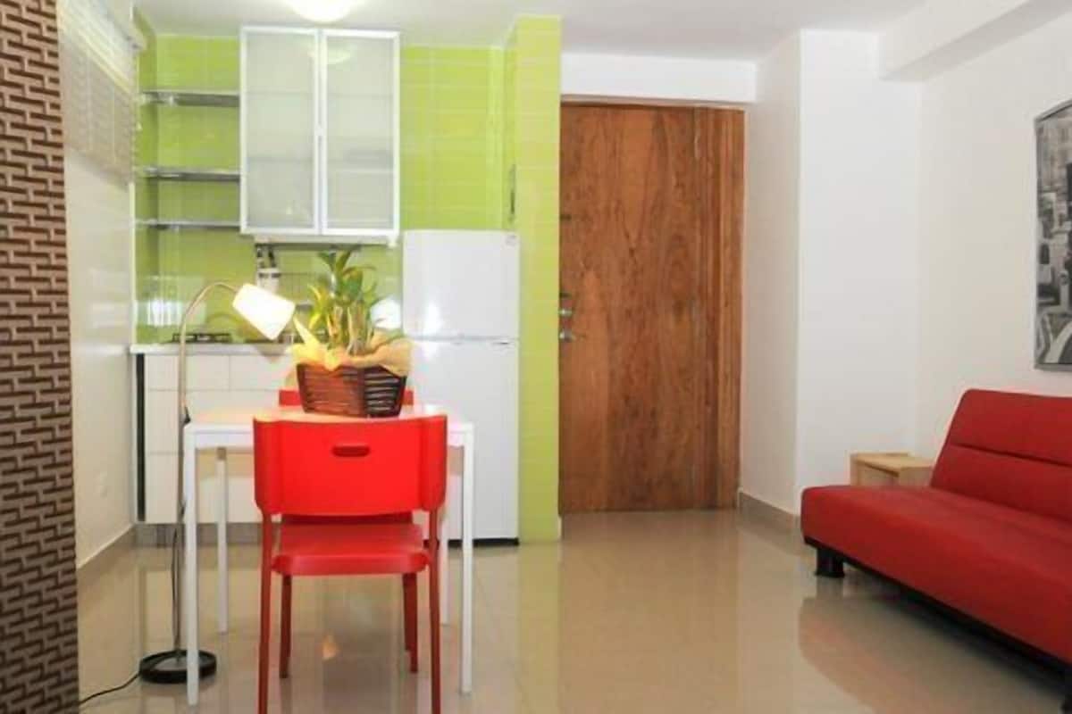 Een ruimtelijk ingerichte leefruimte met kitchenette, een rode stoel, een houten deur en een rode bank.