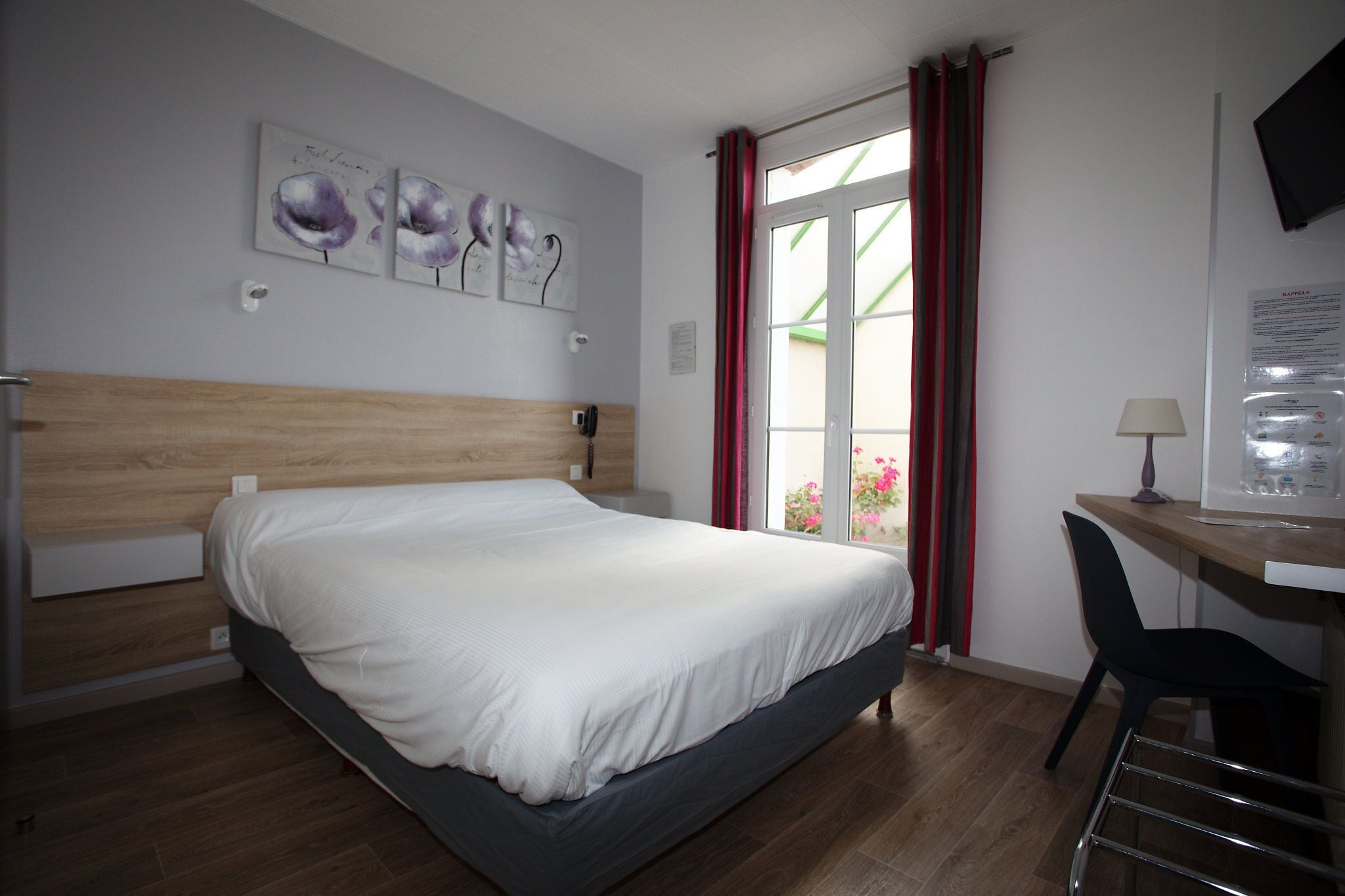 A hotel room with a bed, a desk, a chair, and a window with red curtains.