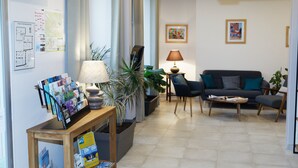 Una sala de estar moderna con un sofá, una mesa de centro y sillas. Hay plantas, una lámpara y un tablón de anuncios con varios papeles.