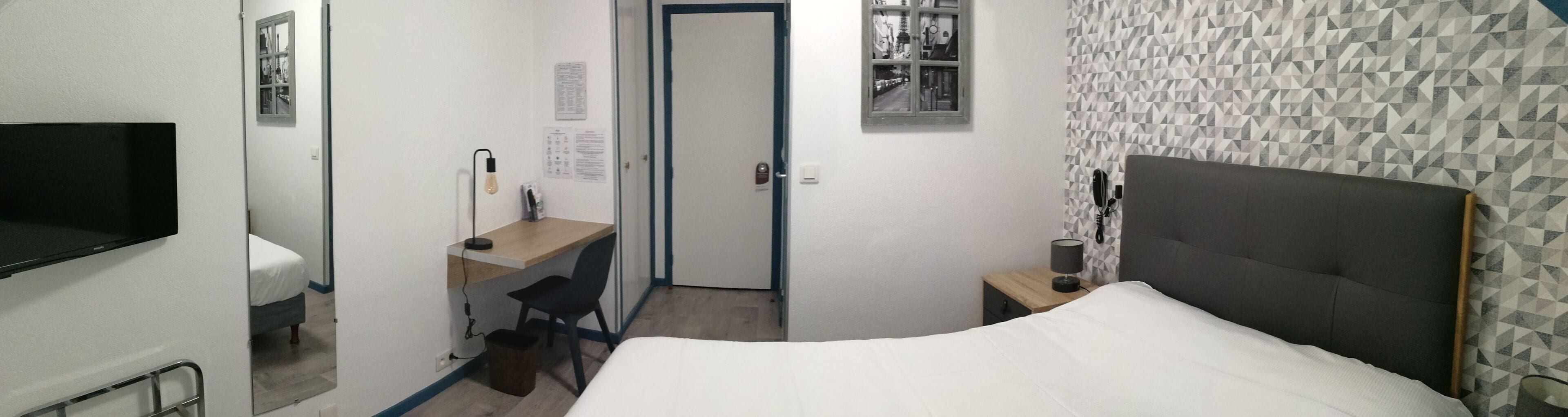 Un dormitorio con cama, escritorio, silla, televisor, mesita de noche con lámpara y puerta que da a otra habitación.