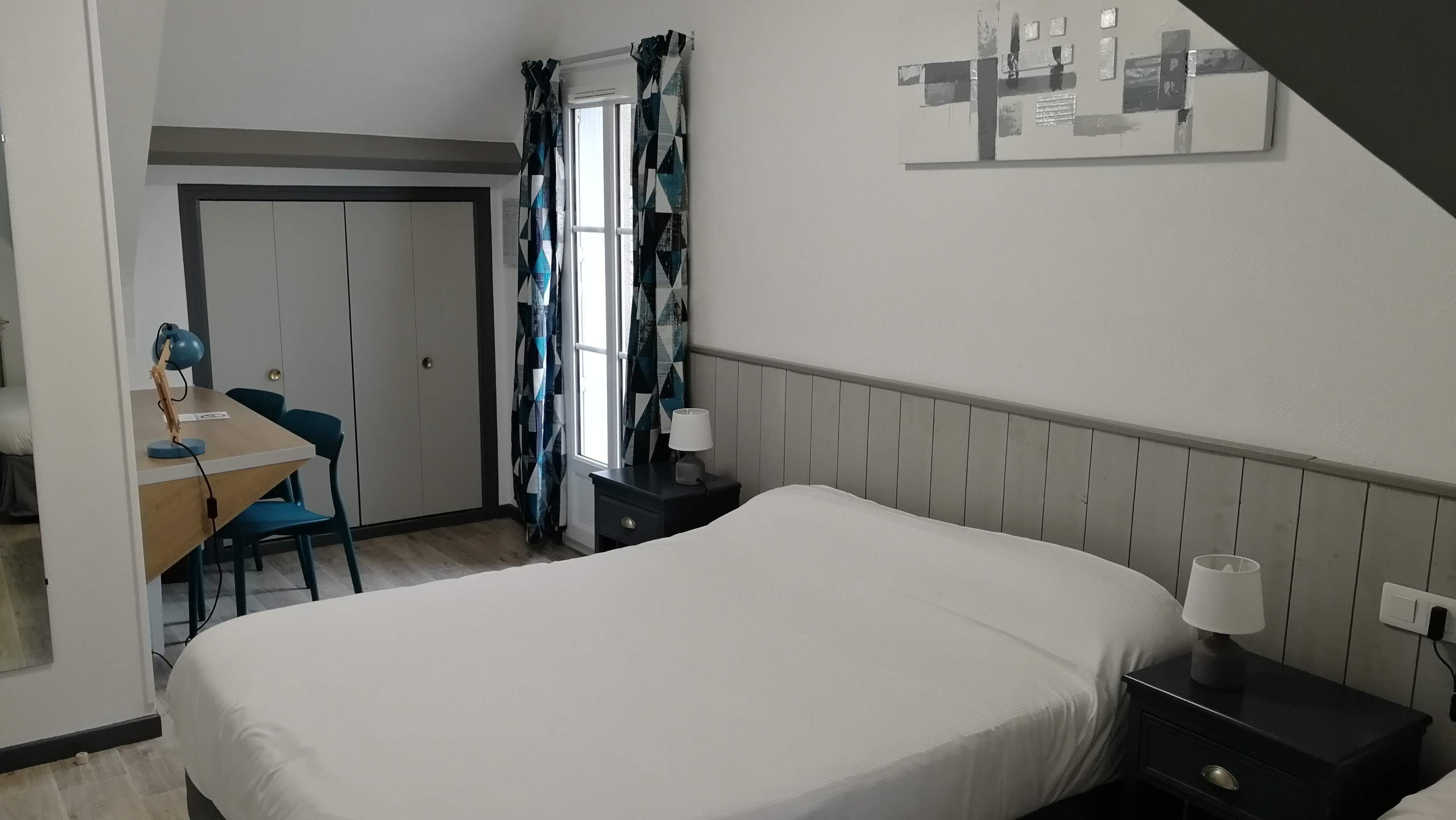 A hotel room with a bed, a desk with a chair, a nightstand, and a window with curtains.