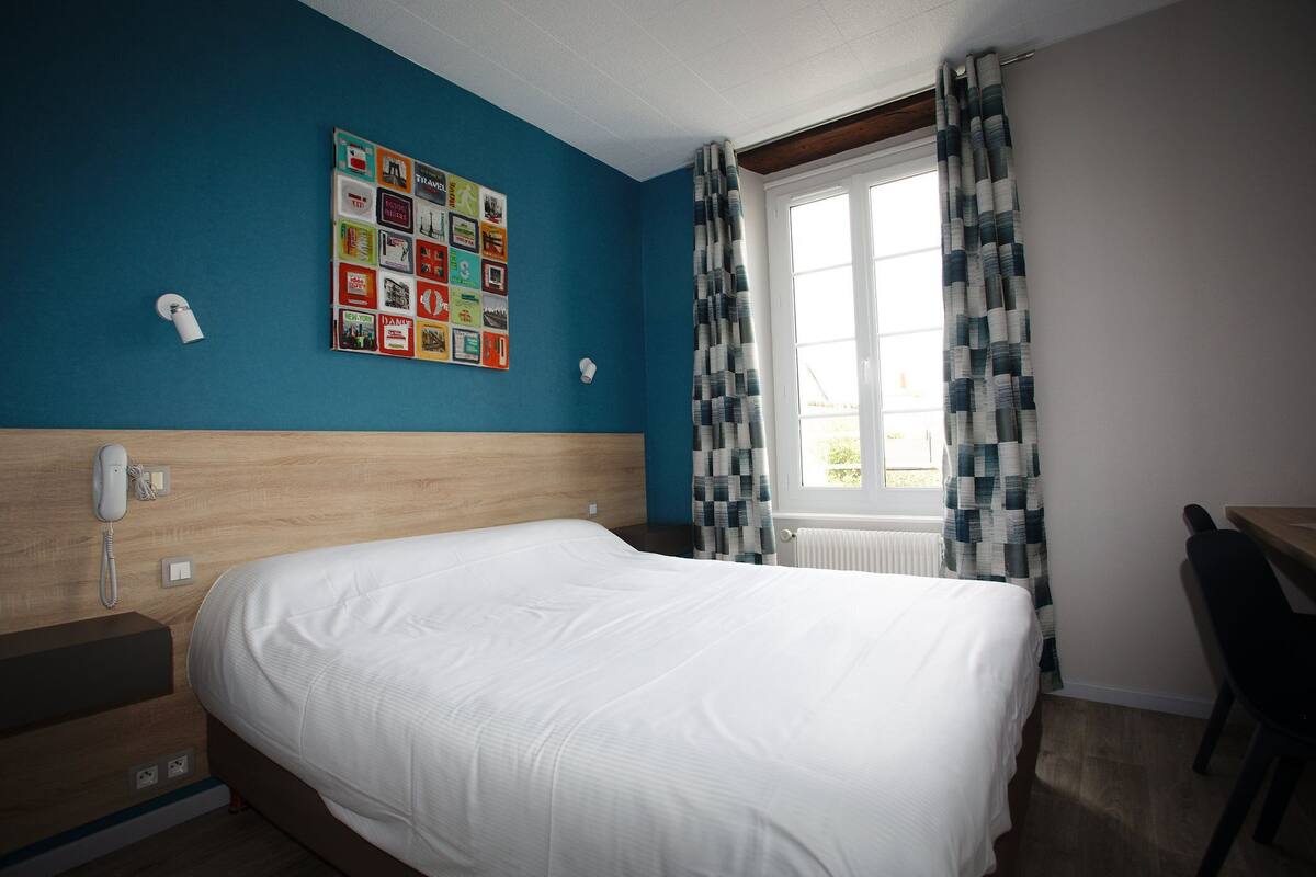 Een hotelkamer met een groot bed, een houten hoofdbord, een aan de muur bevestigd telefoontoestel, een raam met geruite gordijnen en een prikbord met verschillende foto’s.