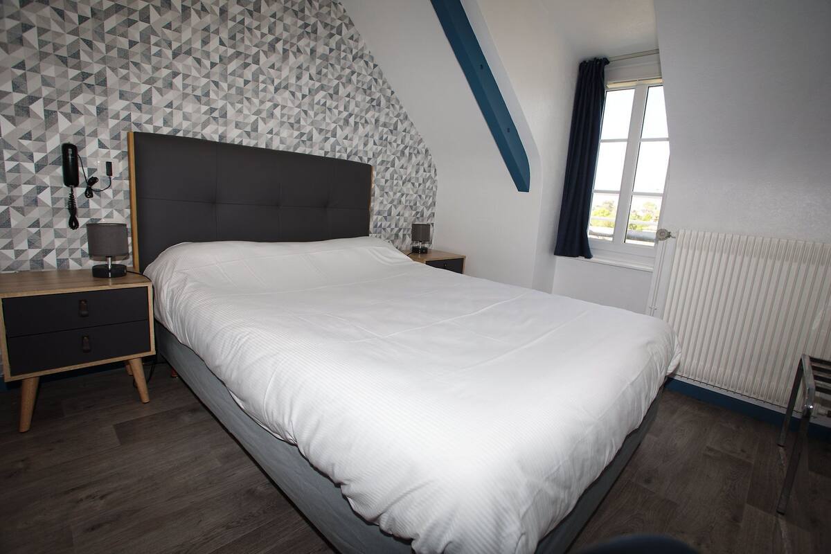Een slaapkamer met een groot bed, een nachtkastje, een lamp, een telefoon en een raam met gordijnen.