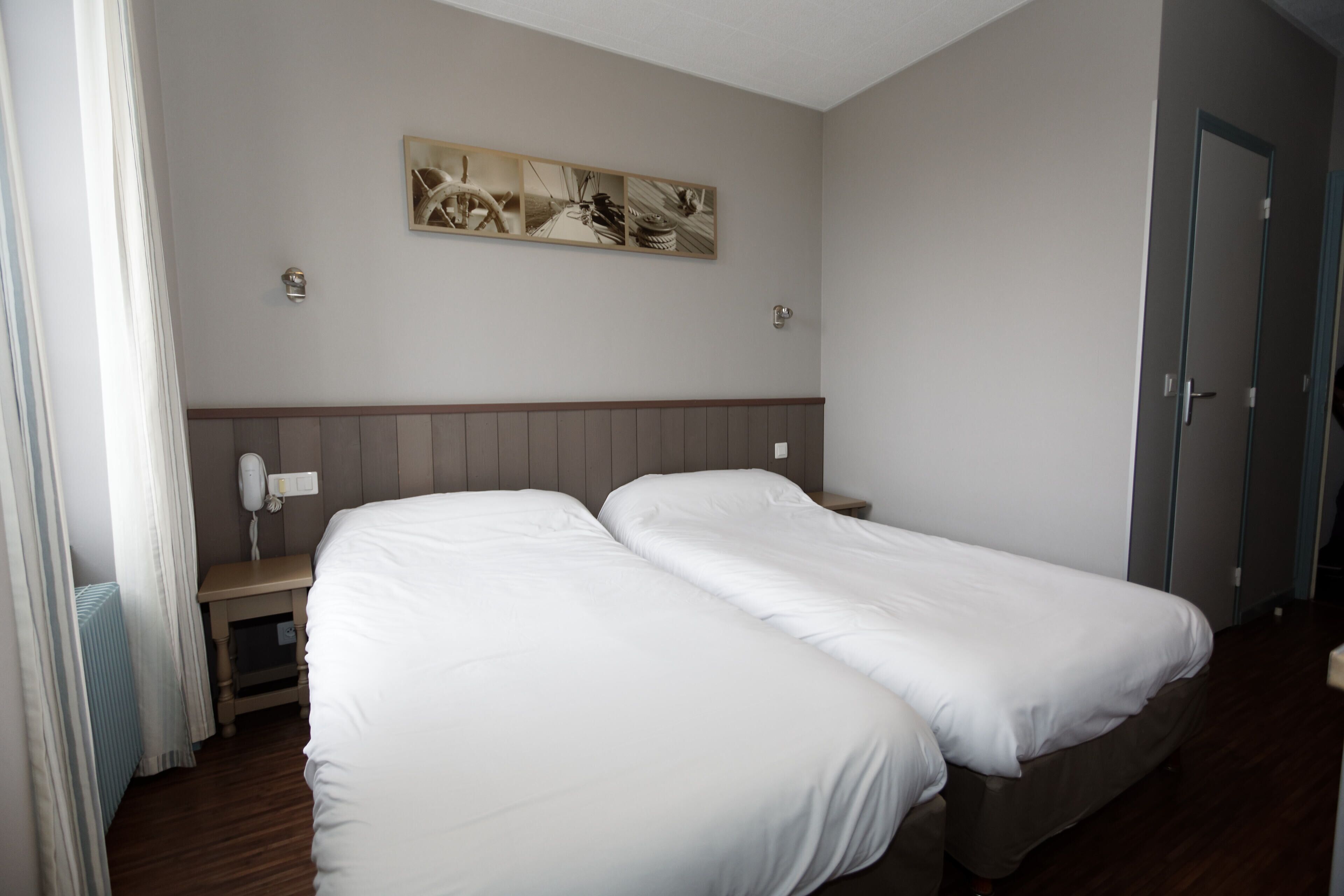 A hotel room with two single beds, a wooden headboard, a bedside table with a lamp, a phone, and a wall-mounted artwork.