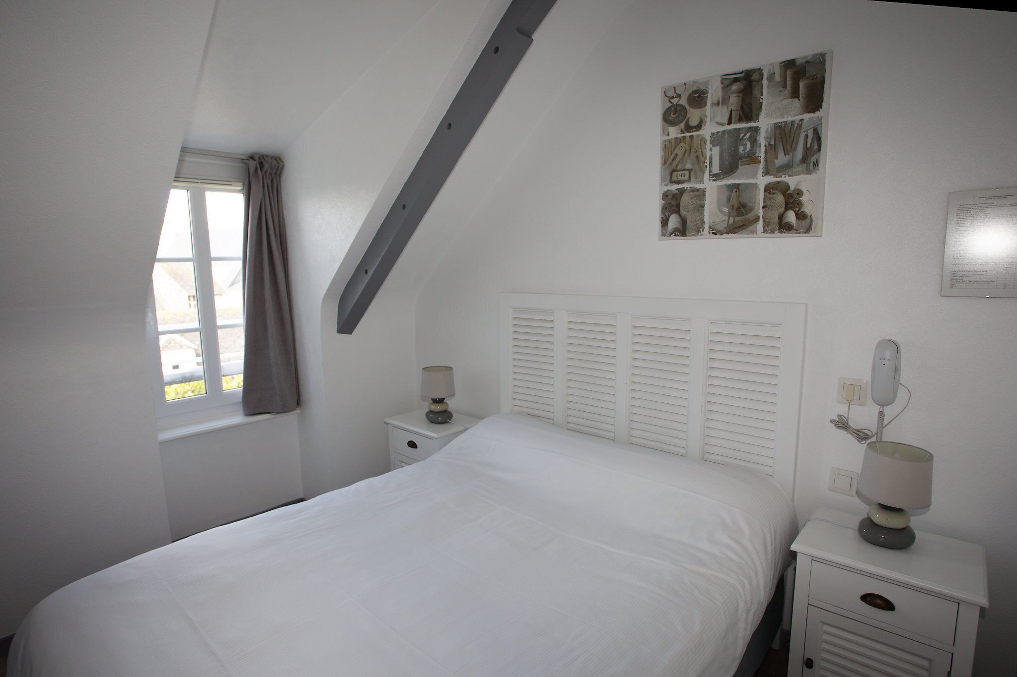 Un dormitorio con cama, dos mesitas de noche, una ventana con cortinas, techo inclinado y una obra de arte colgada en la pared.