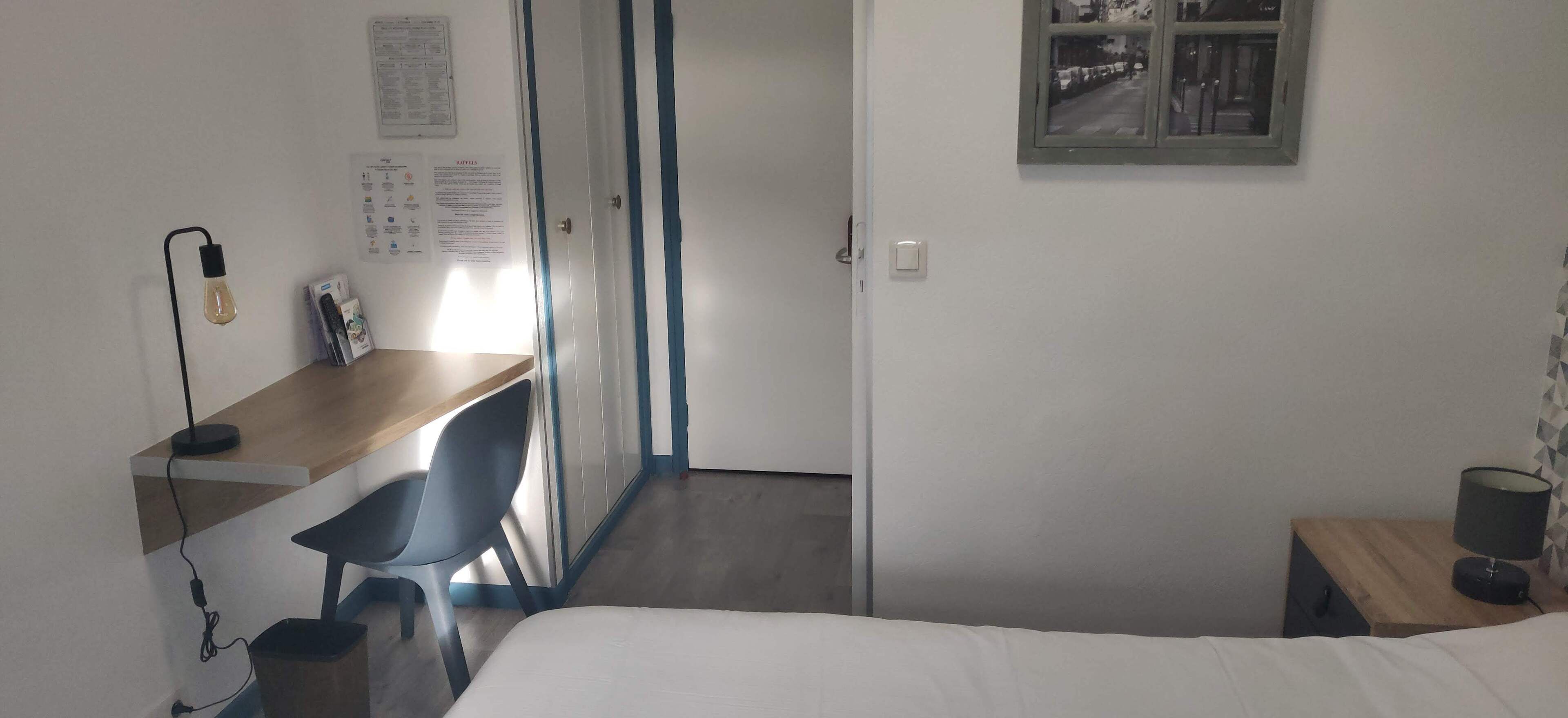 Een slaapkamer met een bed, een bureau met stoel, een lamp en een raam met uitzicht op een straat.