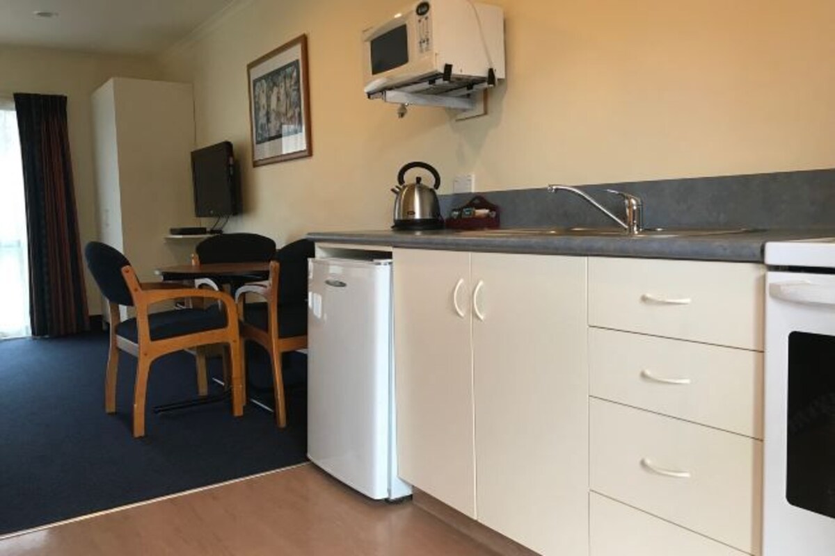 A compact kitchen with a microwave, kettle, and a small refrigerator. There is a dining area with a chair and a table.
