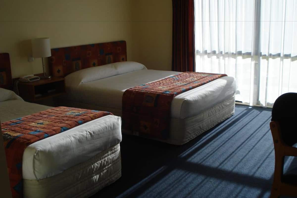 Quarto de hotel com duas camas, uma mesa de cabeceira, um abajur e vista para o exterior através de cortinas translúcidas.