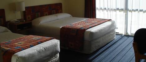 A hotel room with two beds, a nightstand, a lamp, and a view of the outdoors through sheer curtains.