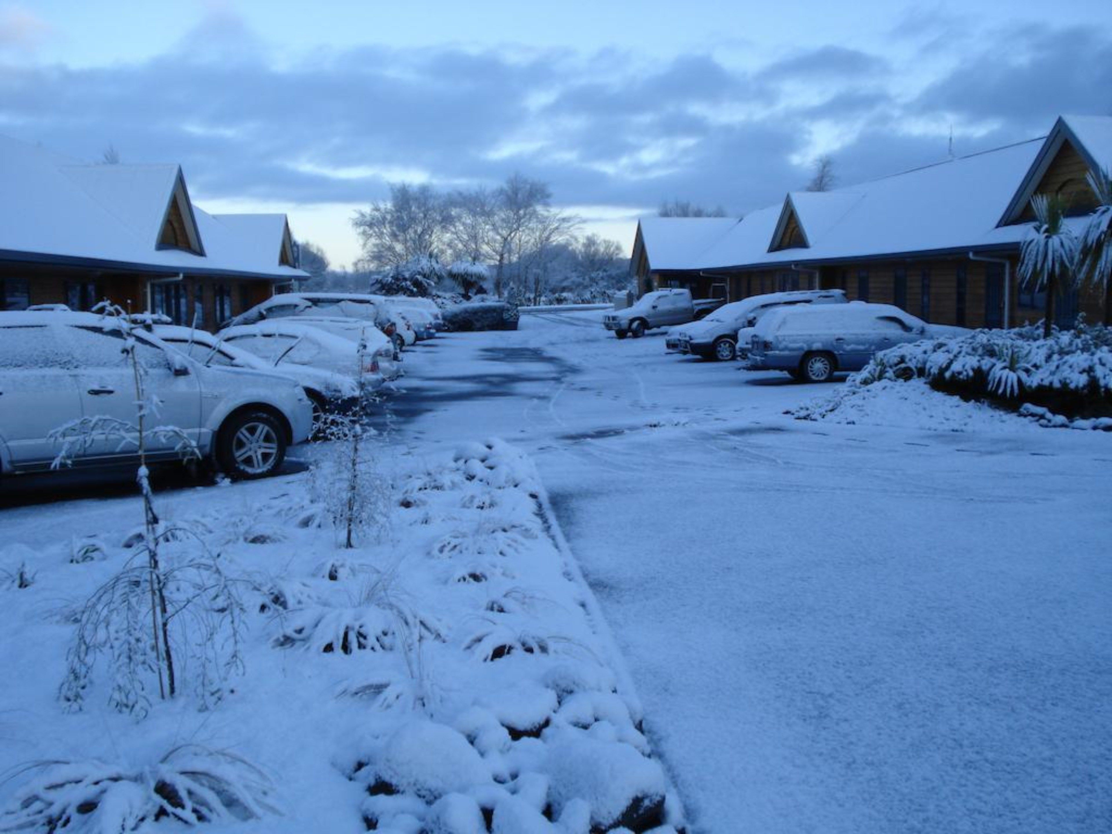 A snow-covered parking lot with several cars parked in front of wooden cabins.