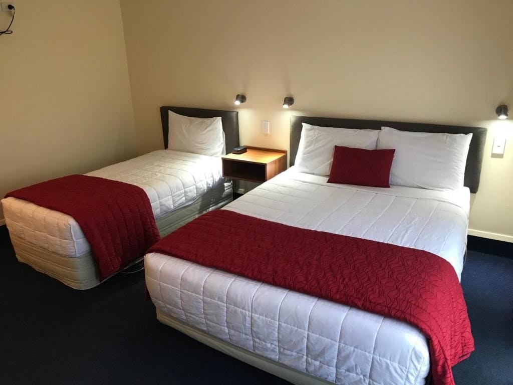 A hotel room with two beds, each with a red blanket and white bedding. There is a nightstand with a lamp between the beds.