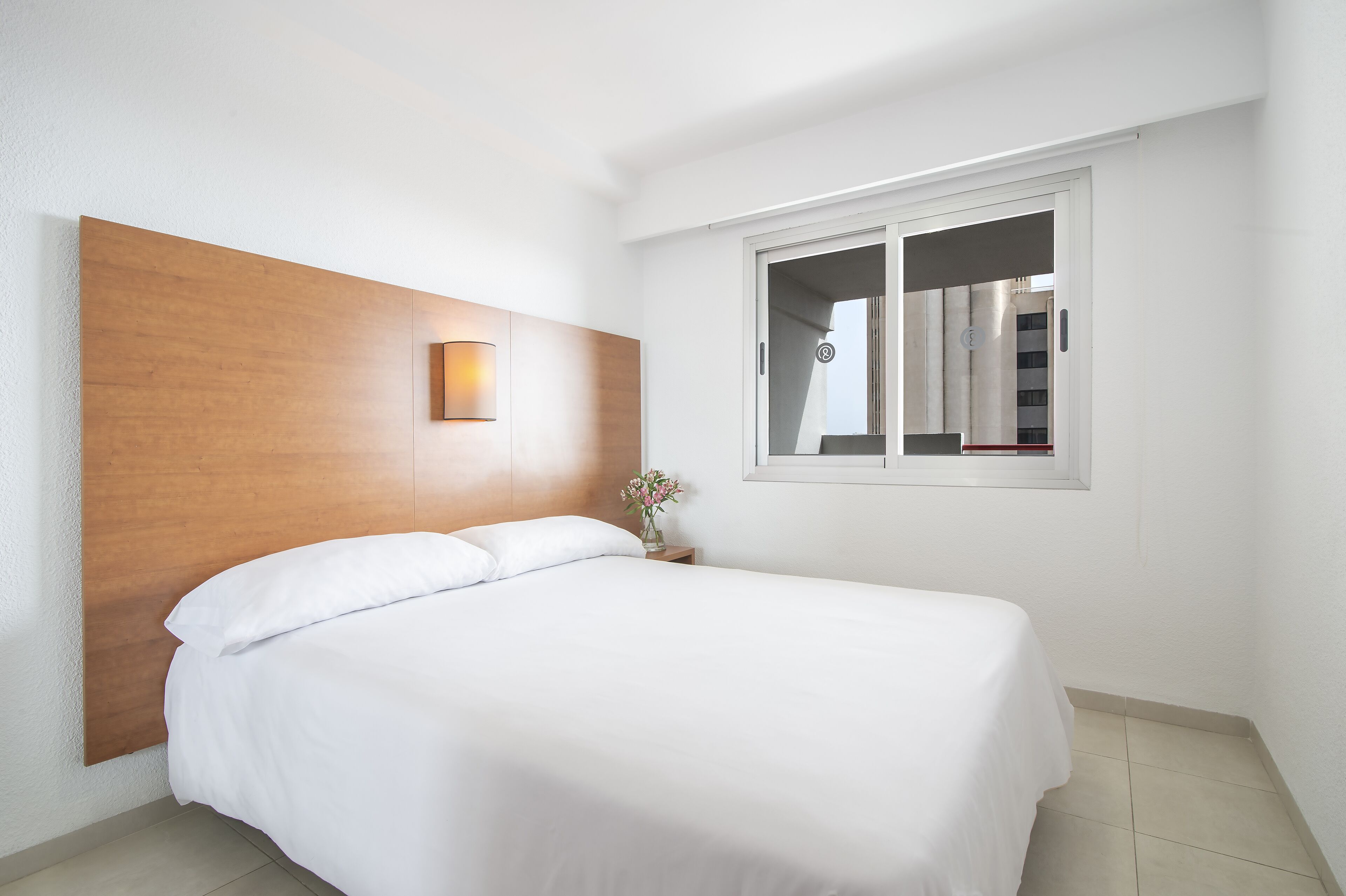 A neatly made bed in a hotel room with a wooden headboard, a small bedside table with a vase, and a window with a view of a building.