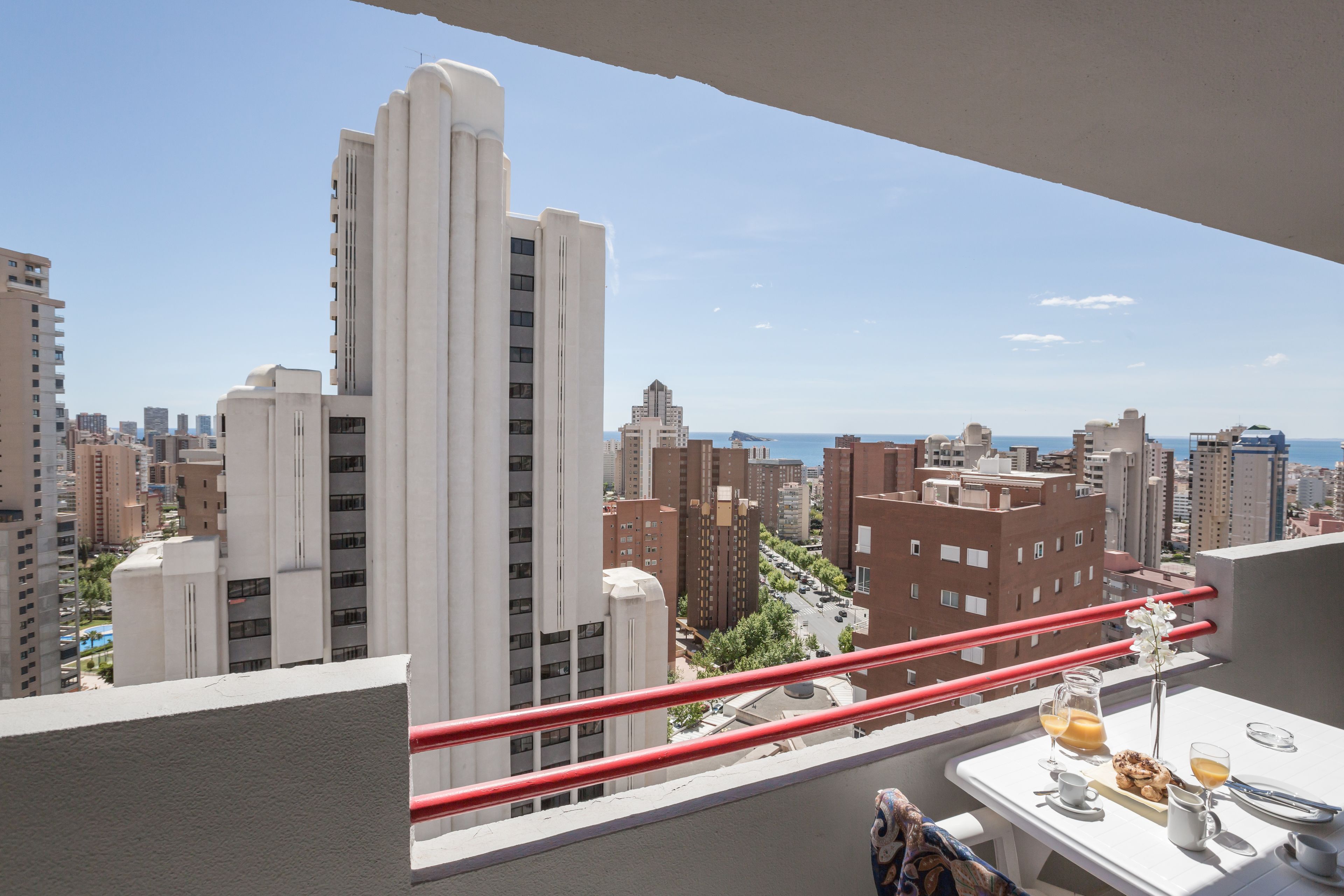 A balcony with a table set for breakfast, overlooking a cityscape with tall buildings and a clear sky.