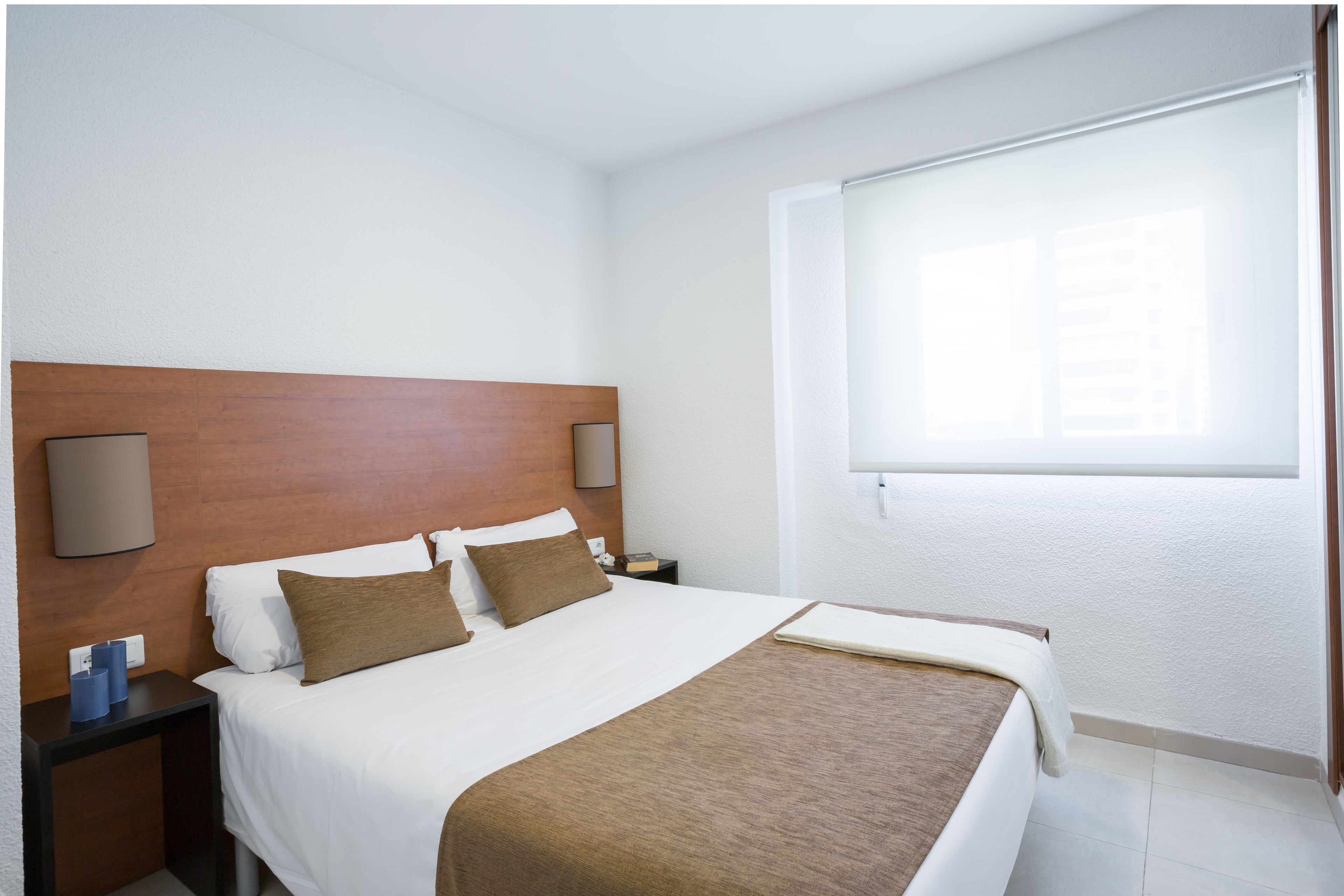 Ein Hotelzimmer mit einem hölzernen Kopfteil, einem großen Bett mit weißer und brauner Bettwäsche, einem Nachttisch mit Lampe und einem Fenster mit Jalousien.