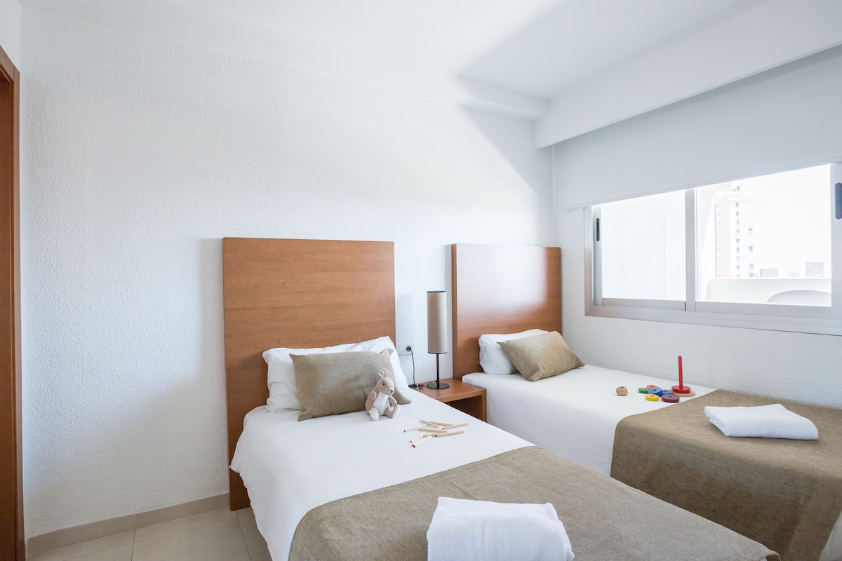 A hotel room with two beds, a bedside table with a lamp, and a window with a city view.