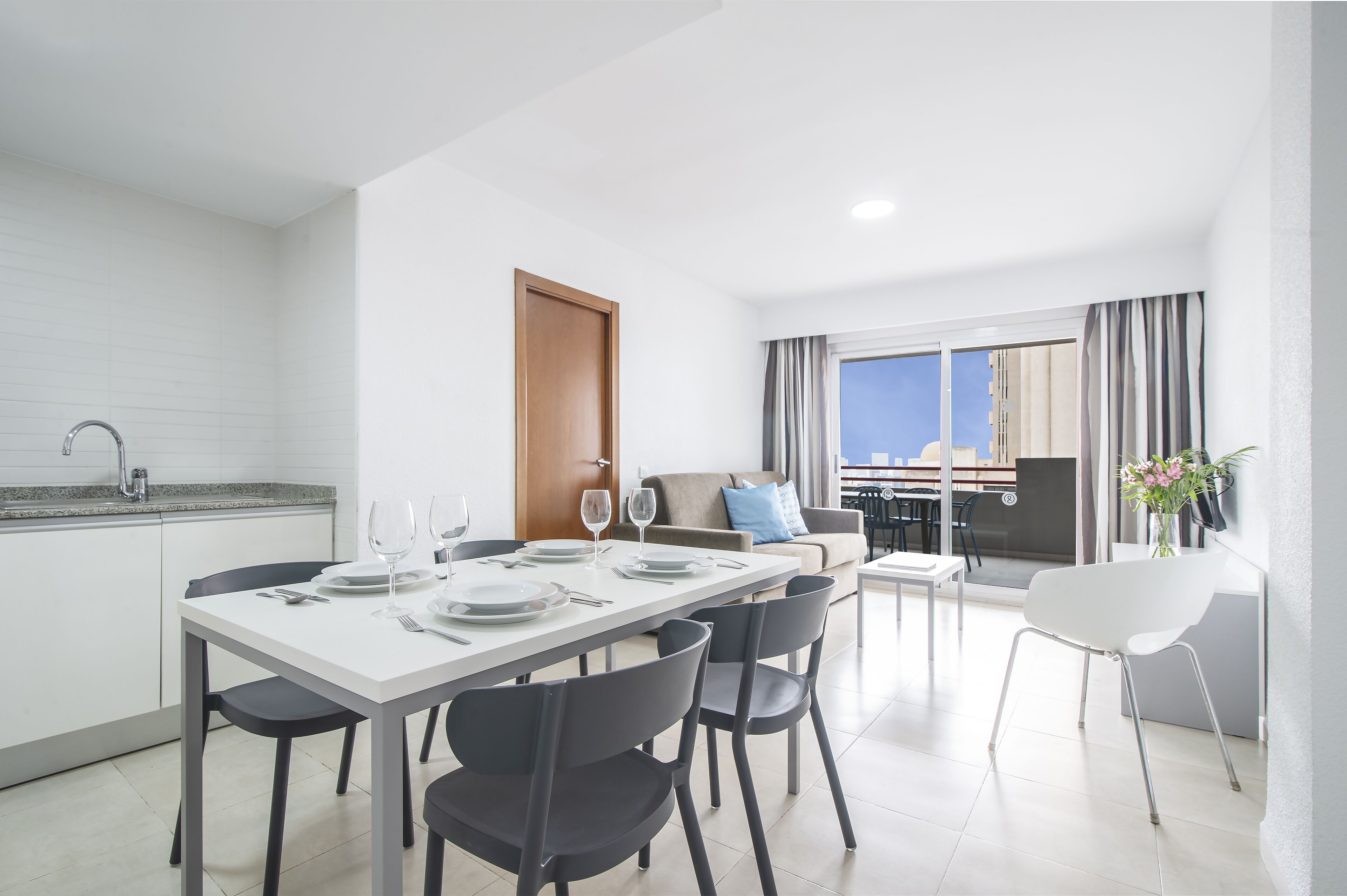 A modern kitchen with white cabinets, a dining area with a white table and black chairs, a sofa with a blue pillow, and a view of the city through large windows.