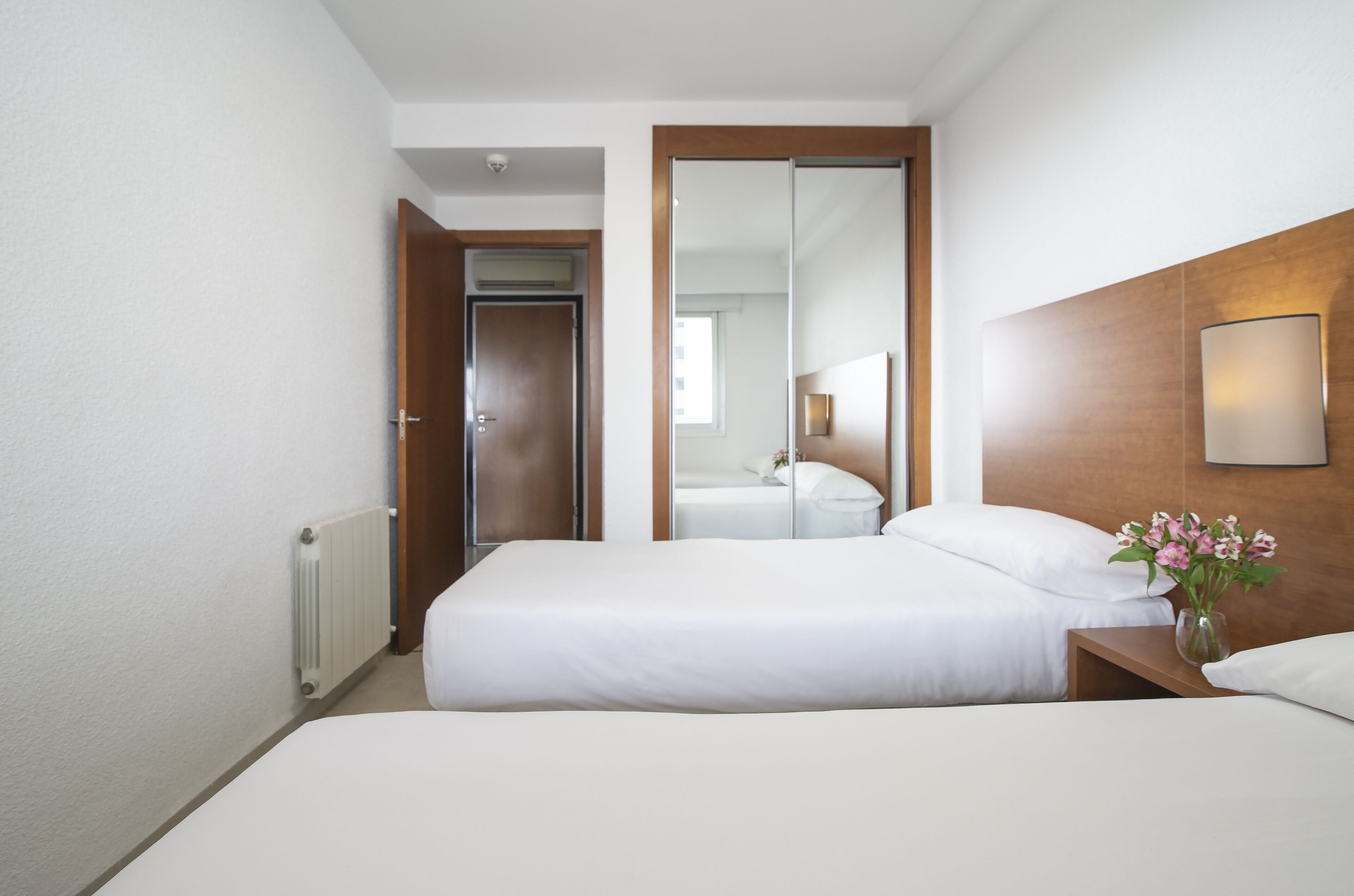 Habitación de hotel con dos camas, cabecero de madera, mesita de noche con un florero y un espejo grande.