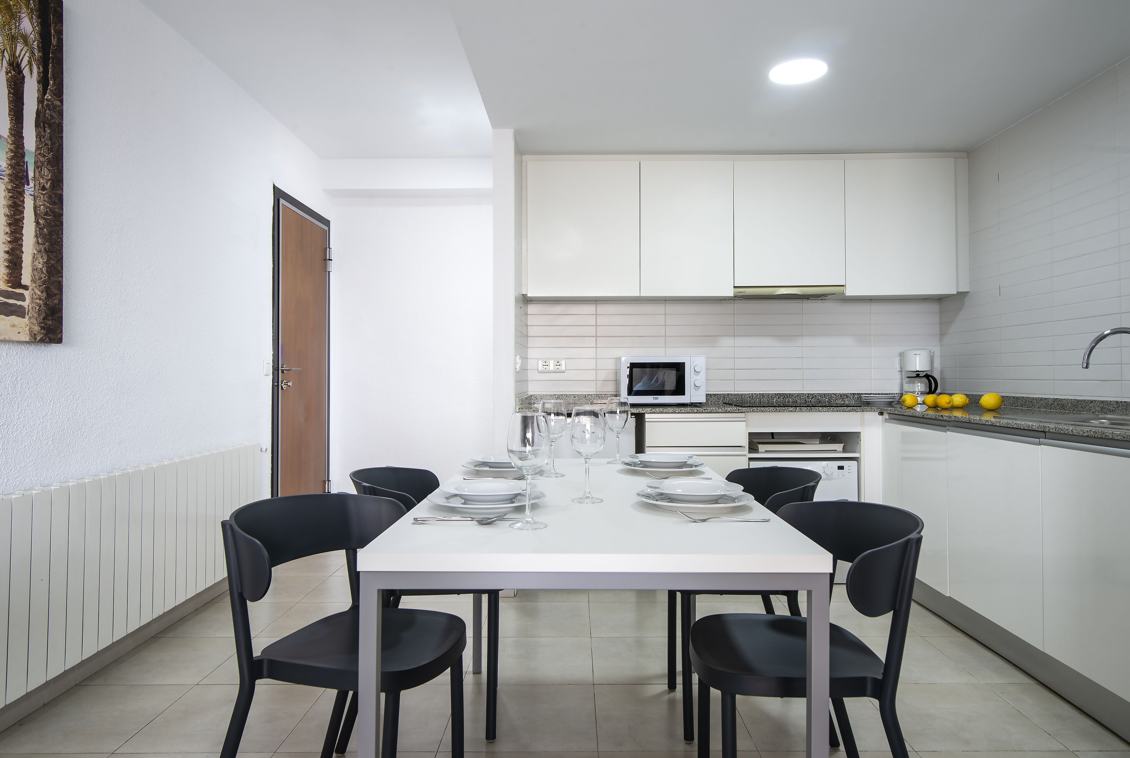 Una cocina moderna con armarios blancos, microondas y cafetera. Hay una mesa de comedor preparada con platos, vasos y cubiertos.