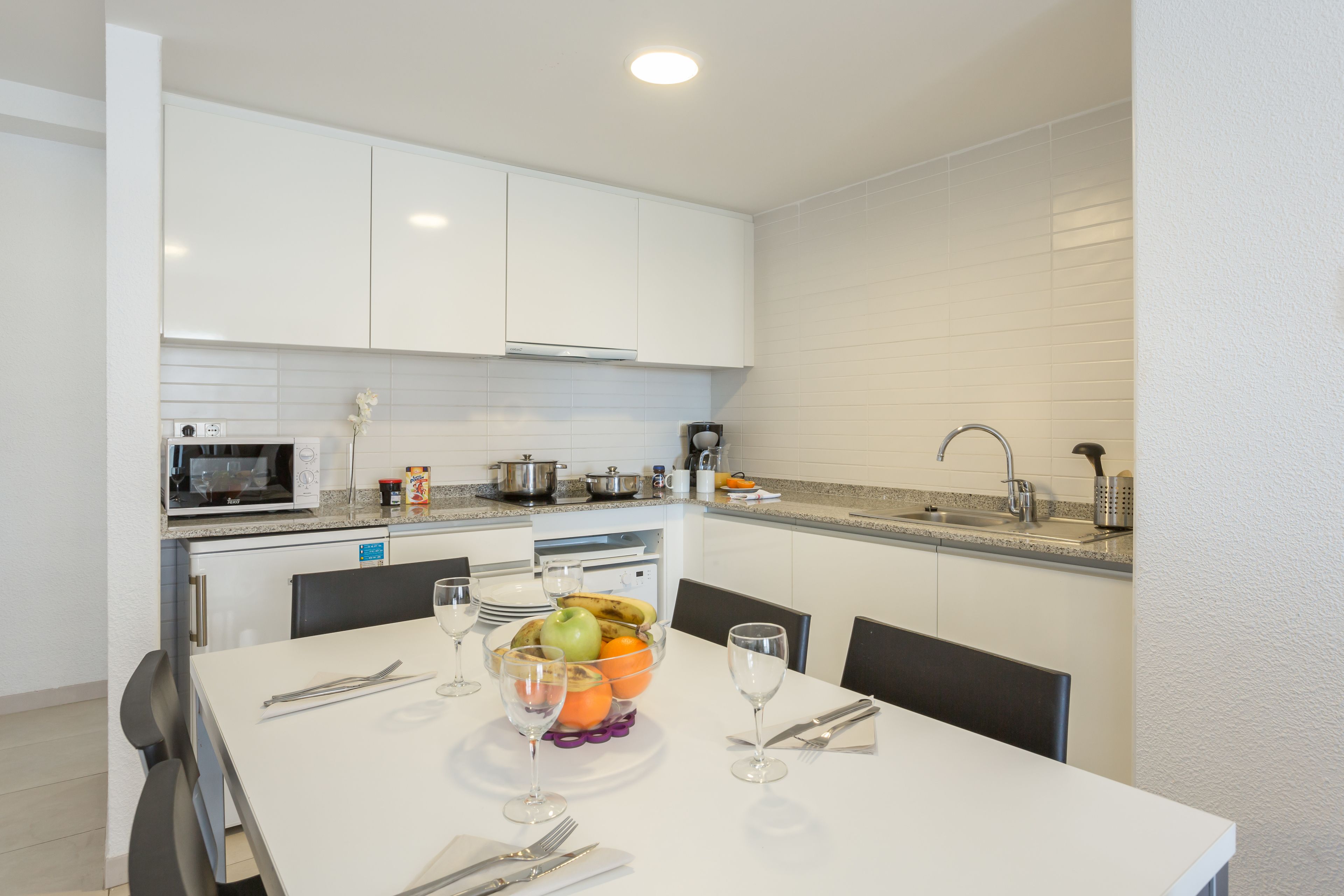 A modern kitchen with white cabinets, a microwave, a sink, and a dining table set with fruit and wine glasses.
