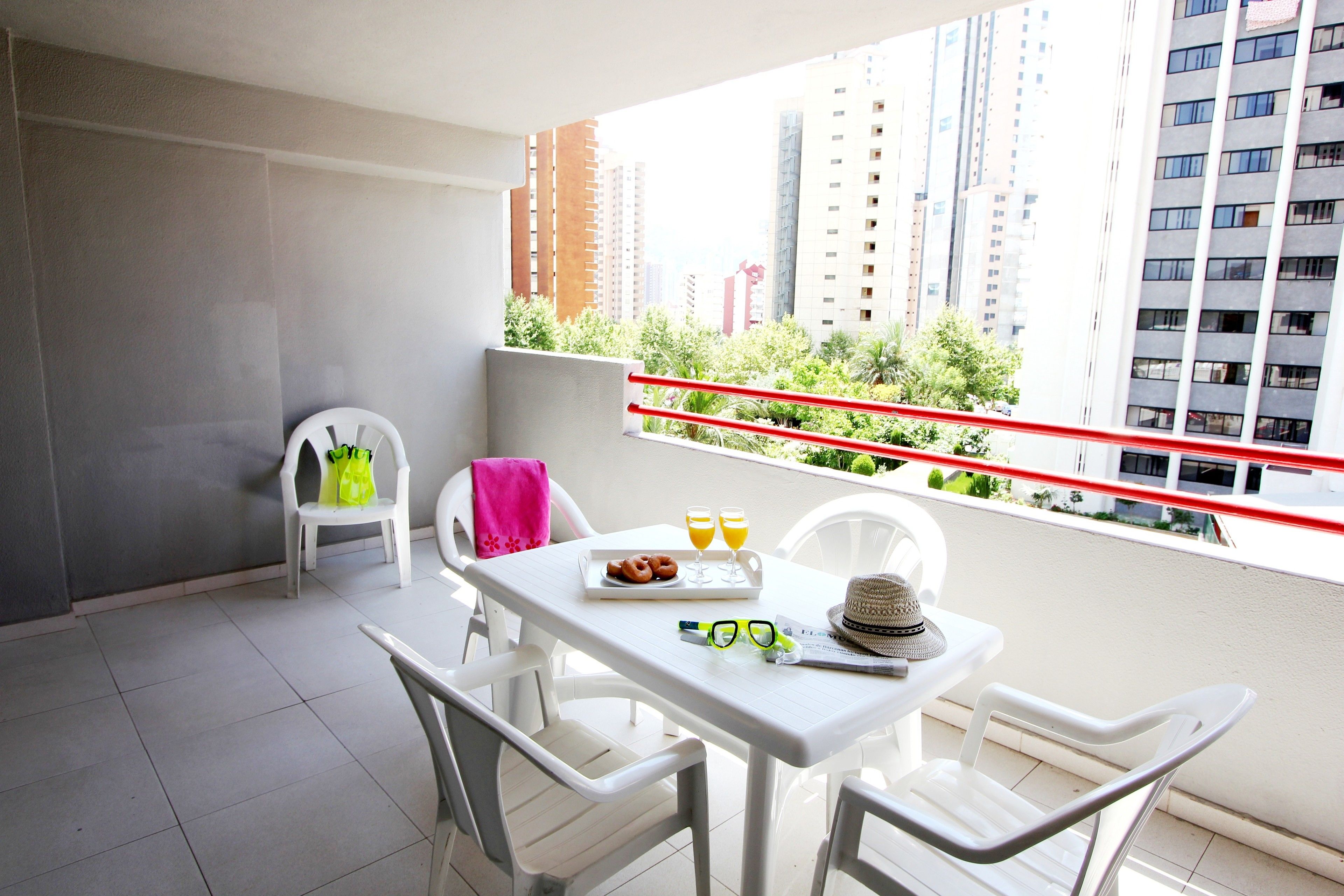 A balcony with a white table, chairs, and a red railing. There are two chairs with a pink towel and a white chair with a green bag. The balcony has a view of tall buildings and greenery.