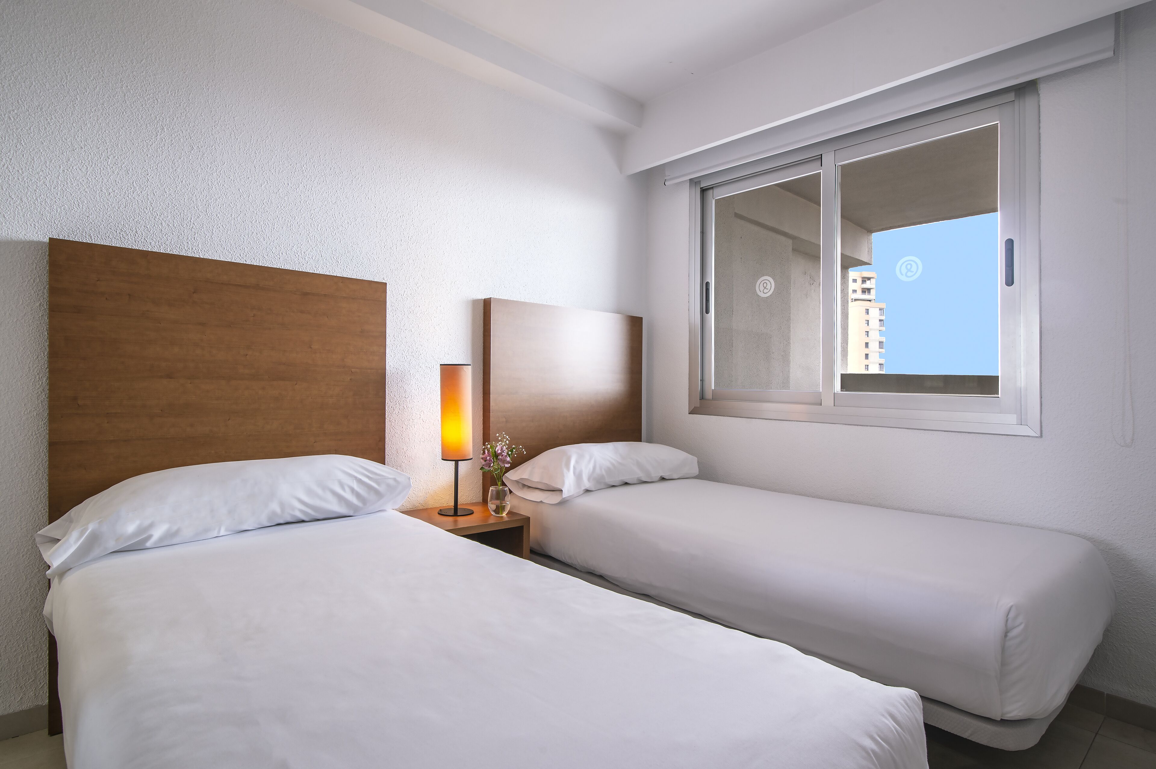 A hotel room with two beds, a wooden headboard, a bedside table with a lamp, and a window with a view of buildings.