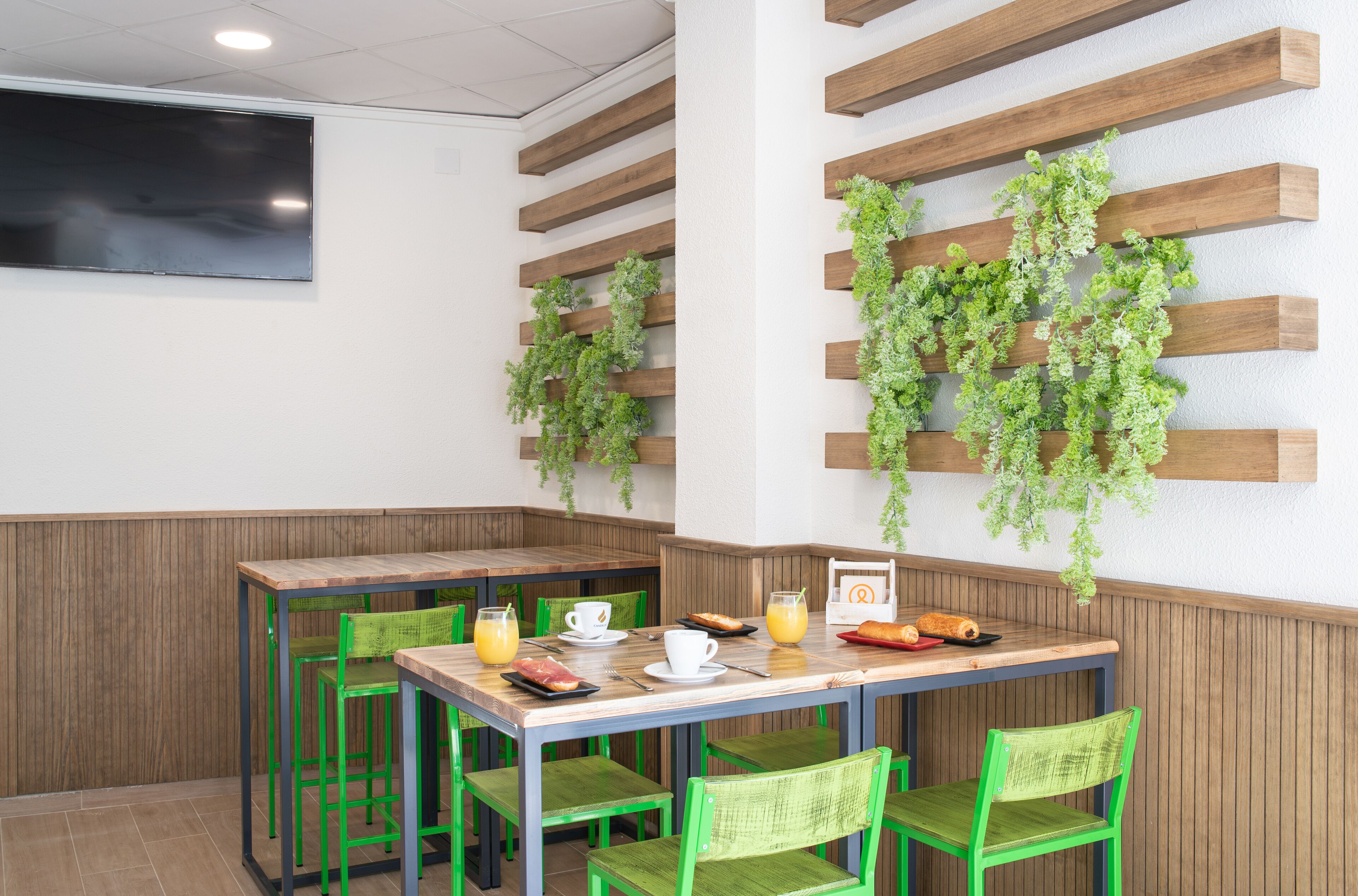 Un comedor con mesas de madera y sillas verdes, un televisor de pantalla plana montado en la pared, y plantas colgantes sujetas a vigas de madera.