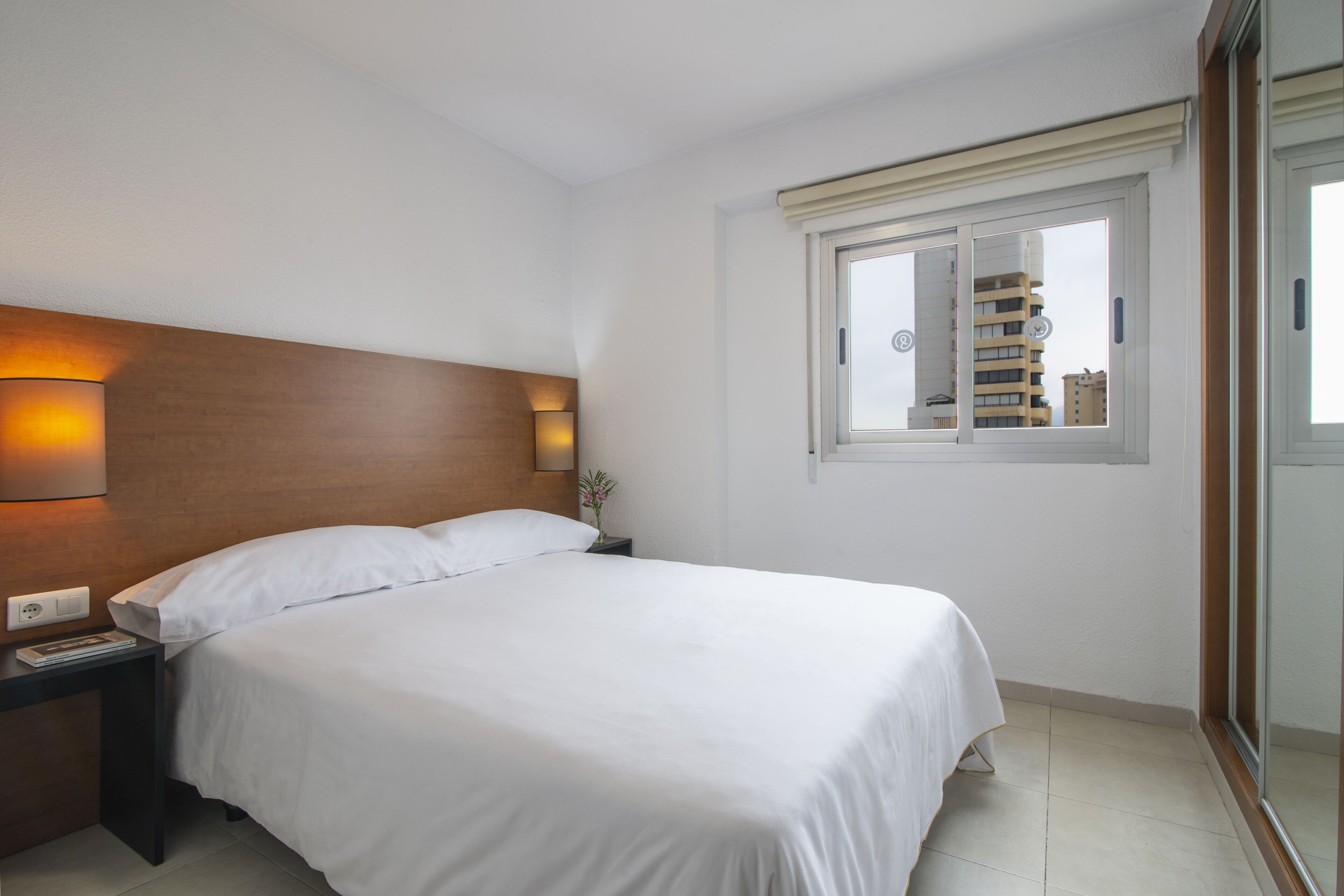 Habitación de hotel con una cama grande, una mesita de noche con lámpara y una ventana con vista a edificios.