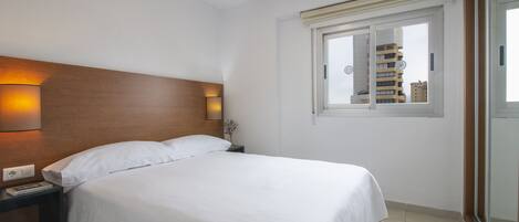Habitación de hotel con una cama grande, una mesita de noche con lámpara y una ventana con vista a edificios.