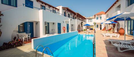 A swimming pool with a blue interior, white walls, and a tiled patio area with lounge chairs and umbrellas.
