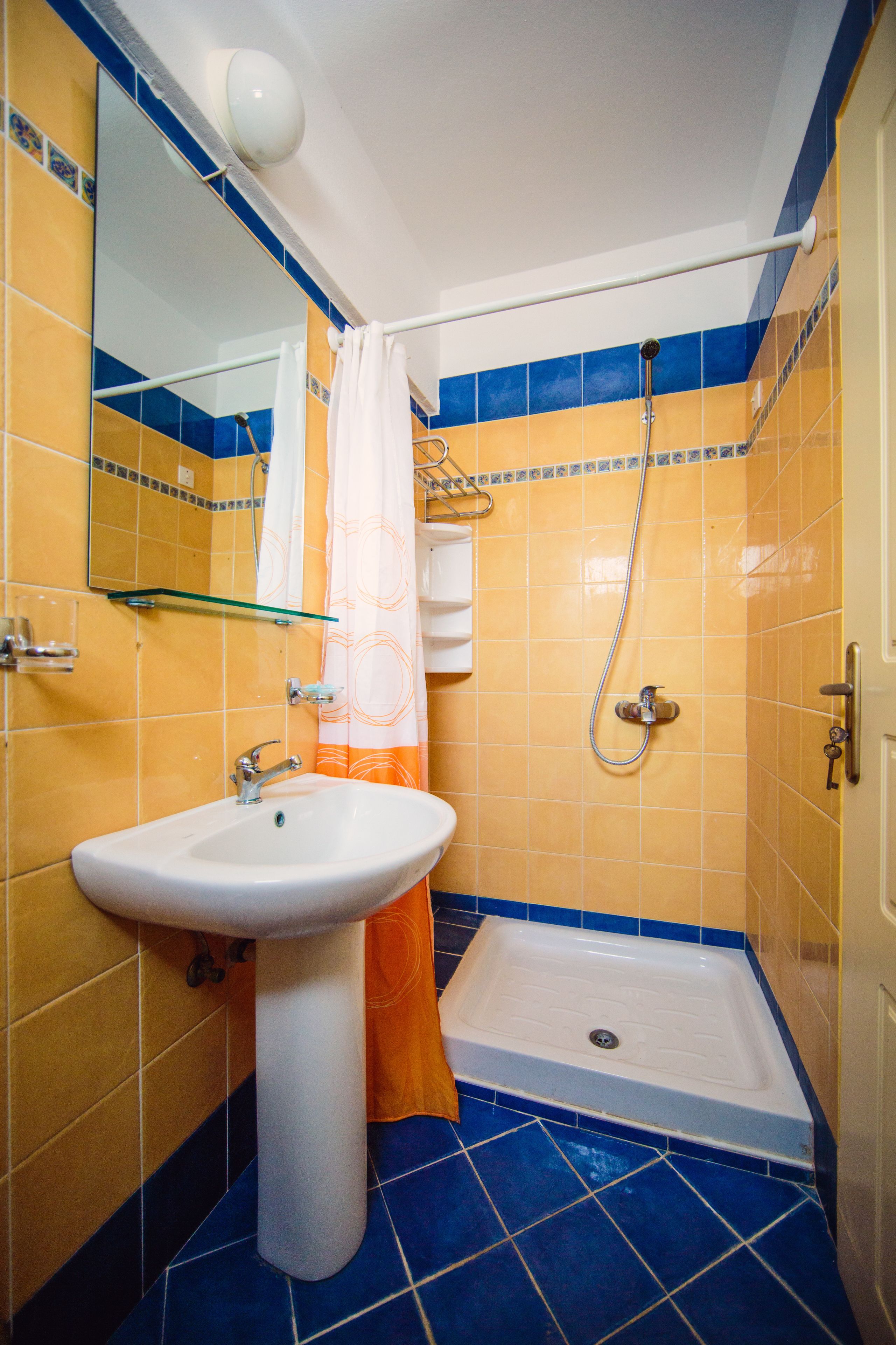 A bathroom with a white sink, a glass shelf, a shower with a handheld showerhead, and a blue-tiled floor.
