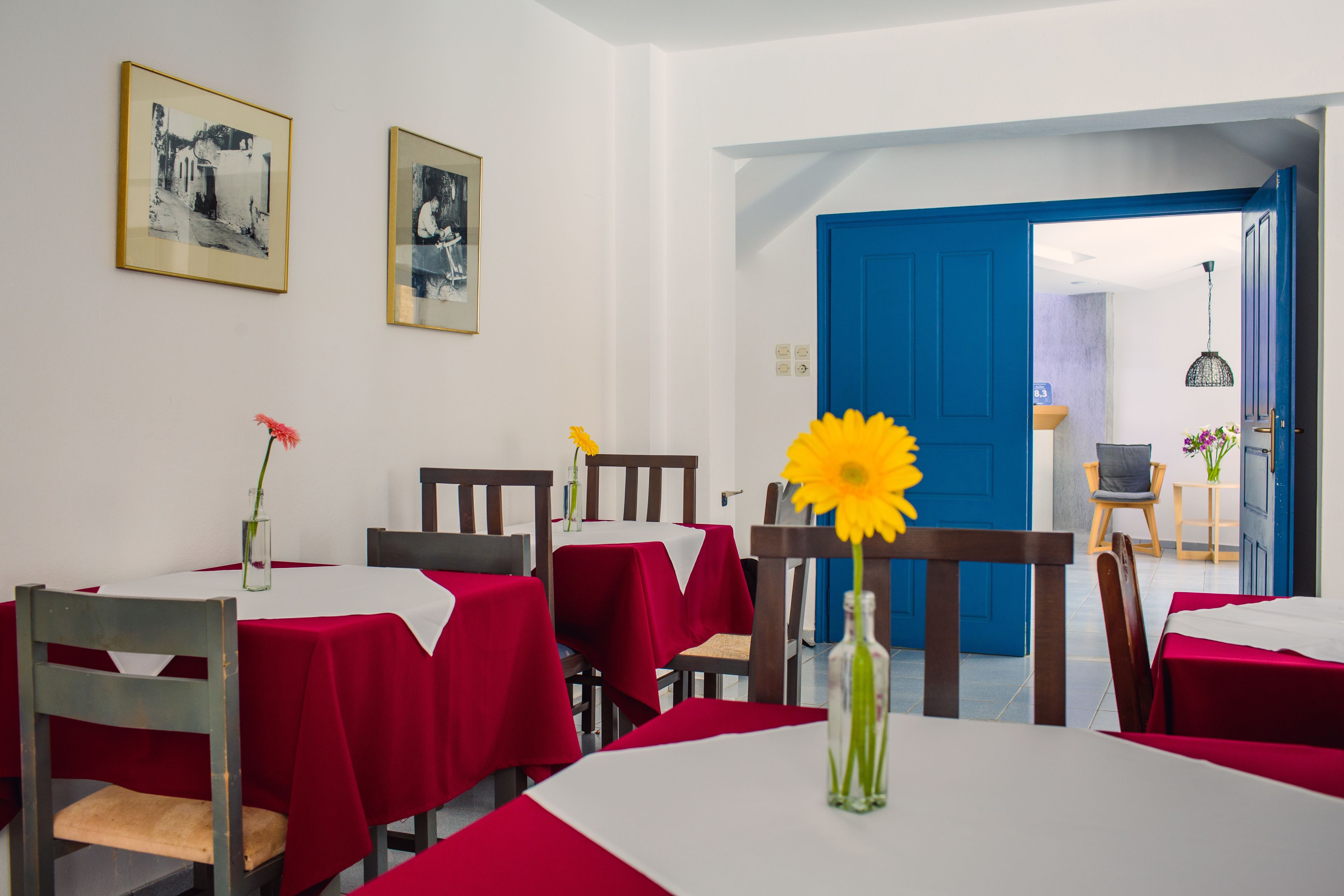 A dining area with tables covered in red tablecloths, wooden chairs, and a vase with a yellow flower.