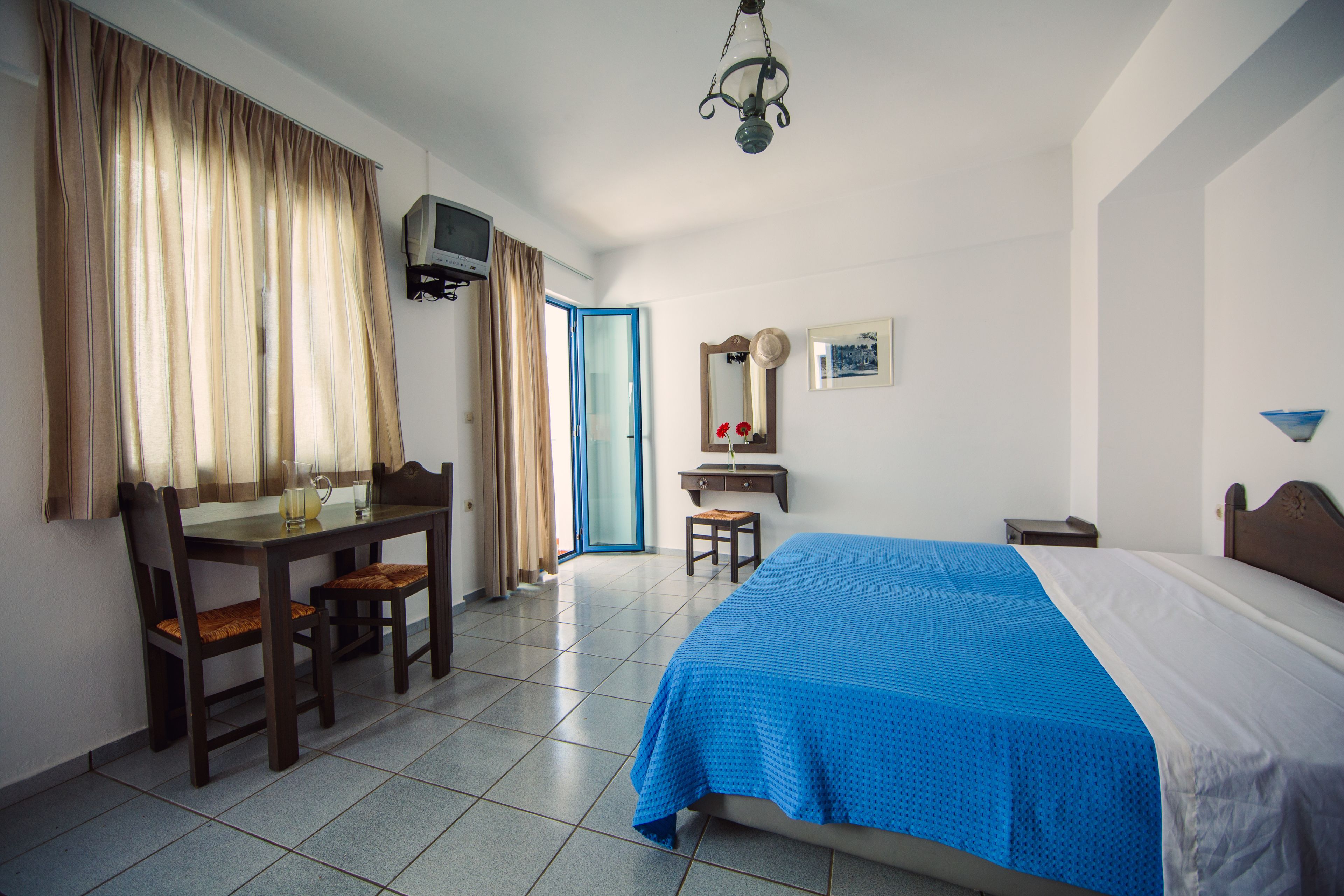 A hotel room with a bed, a desk with chairs, a television, and a door leading to a balcony.