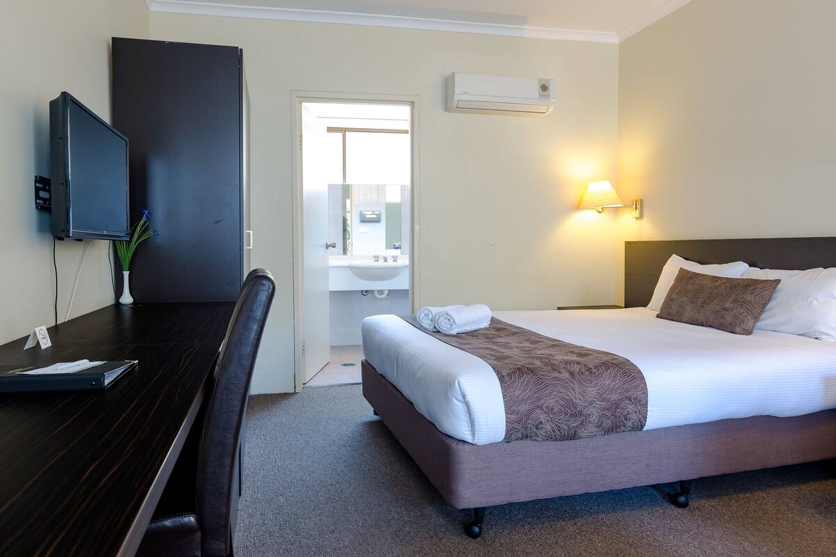 Quarto de hotel com cama, escrivaninha, cadeira, televisão, banheiro e ar-condicionado fixado na parede.