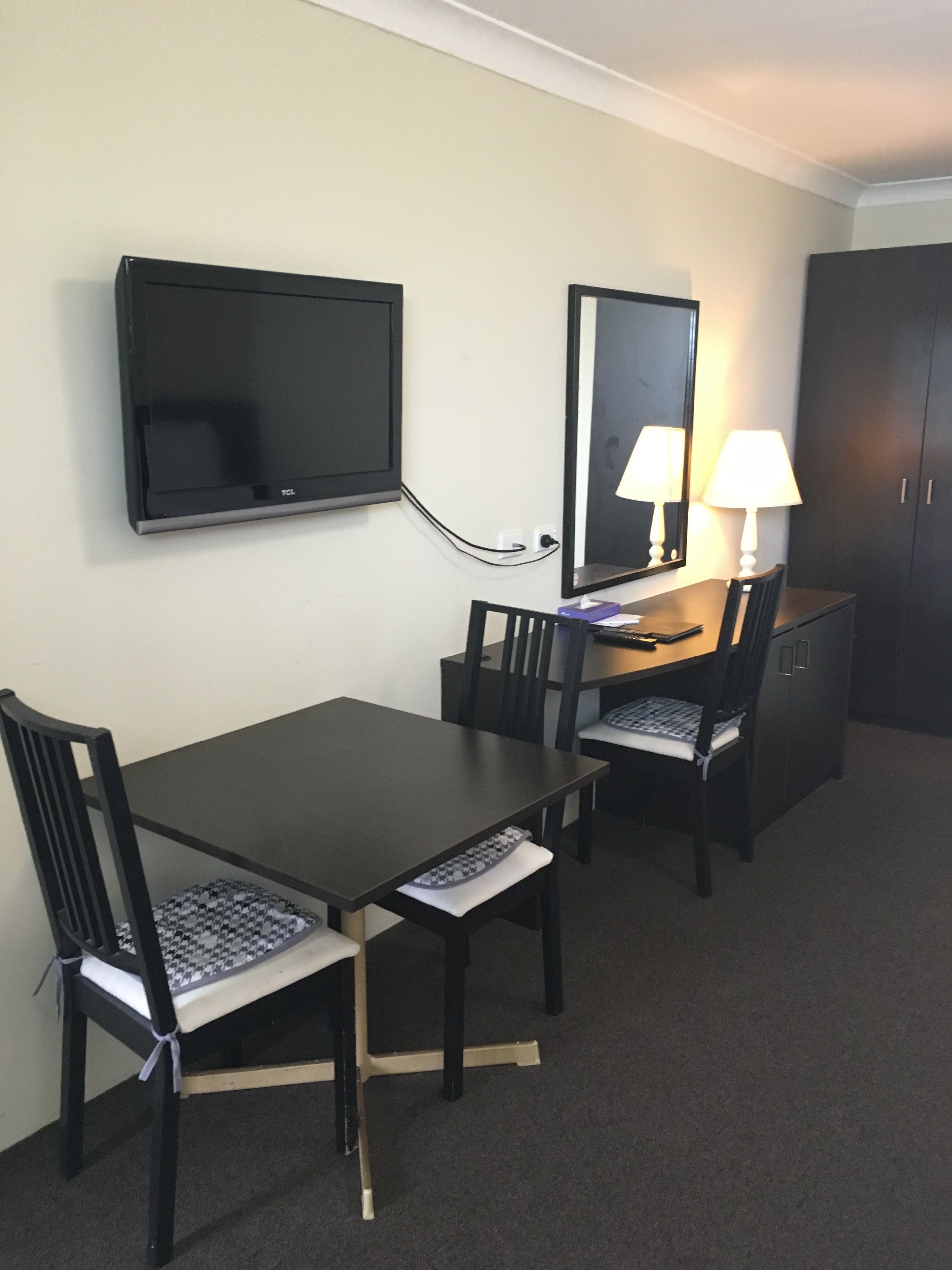 A hotel room with a dark wooden desk, two chairs, a flat-screen TV mounted on the wall, a mirror above the desk, and a lamp on the right side.