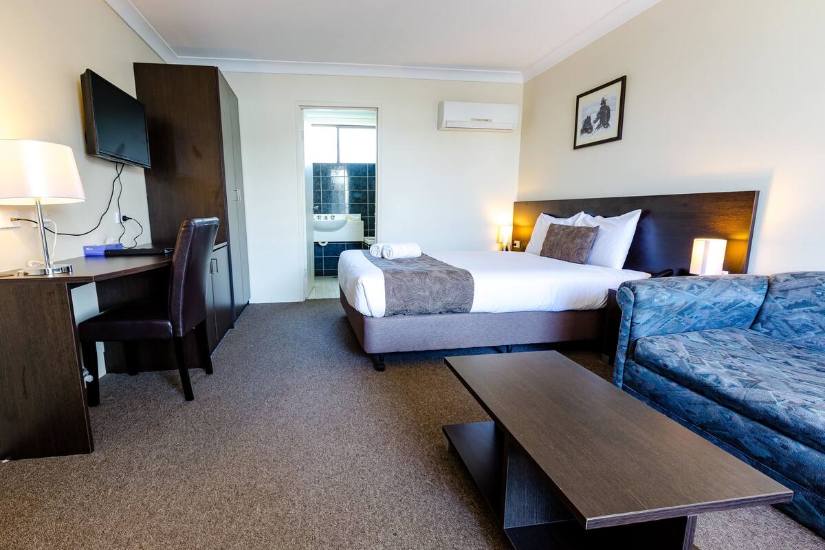 Quarto de hotel com cama, escrivaninha, cadeira, sofá e televisão.