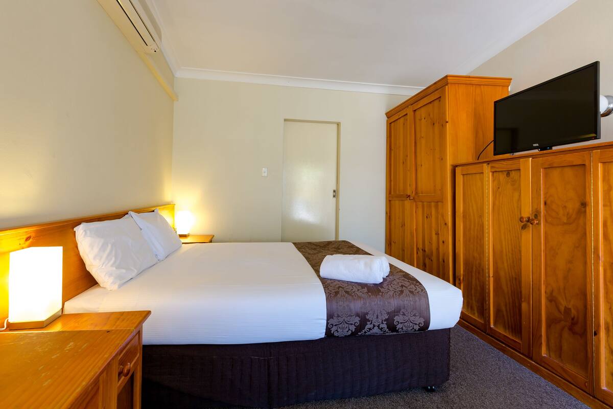 Cama bem arrumada com lençóis brancos e castanhos, mesa de cabeceira de madeira com abajur e guarda-roupa com uma televisão em cima.