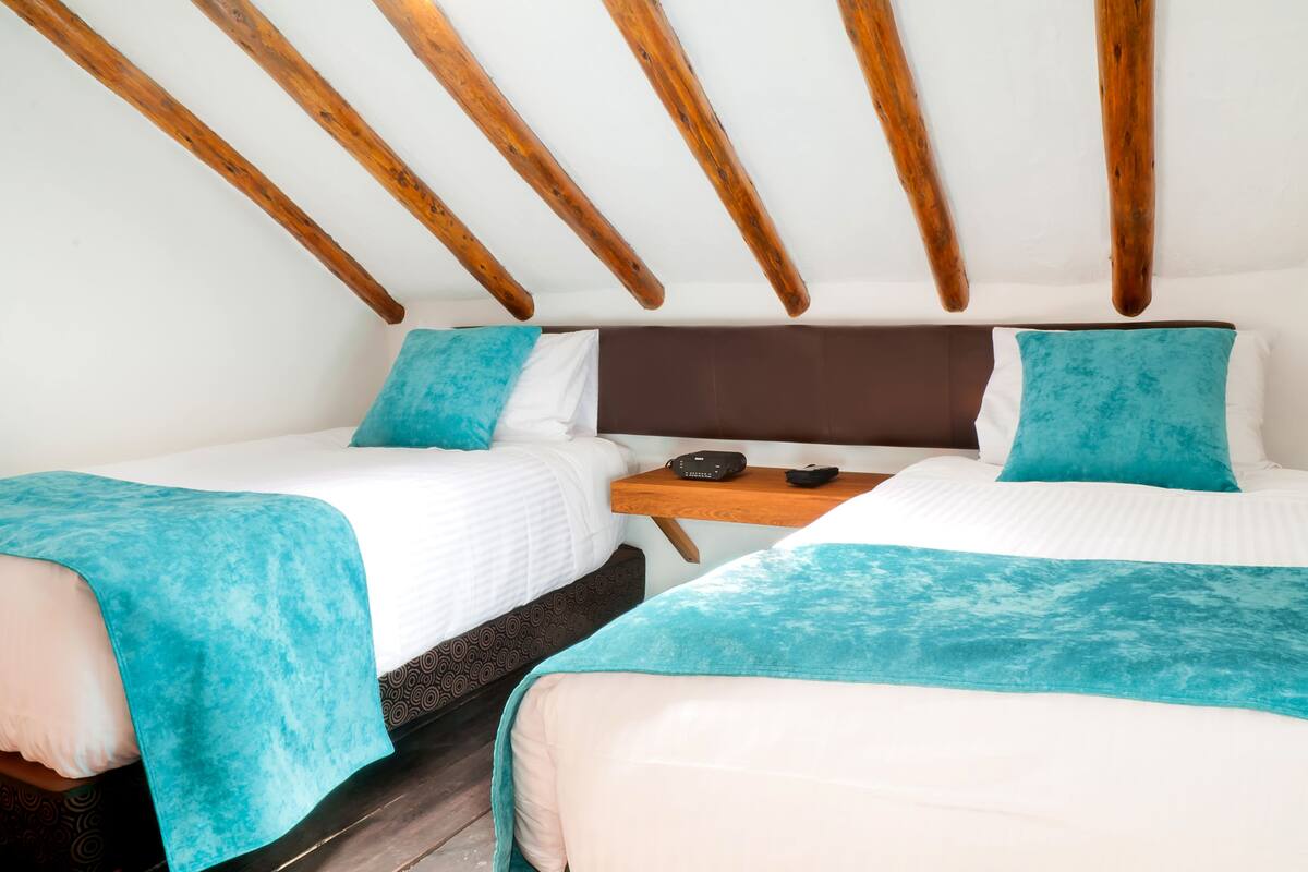 A room with two beds, each with a teal blanket and white bedding. There is a wooden nightstand between the beds with a small table and a remote control.