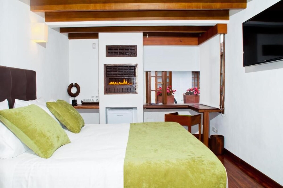 Un dormitorio con cama, almohadas verdes, un escritorio con computadora, chimenea y televisor montado en la pared.