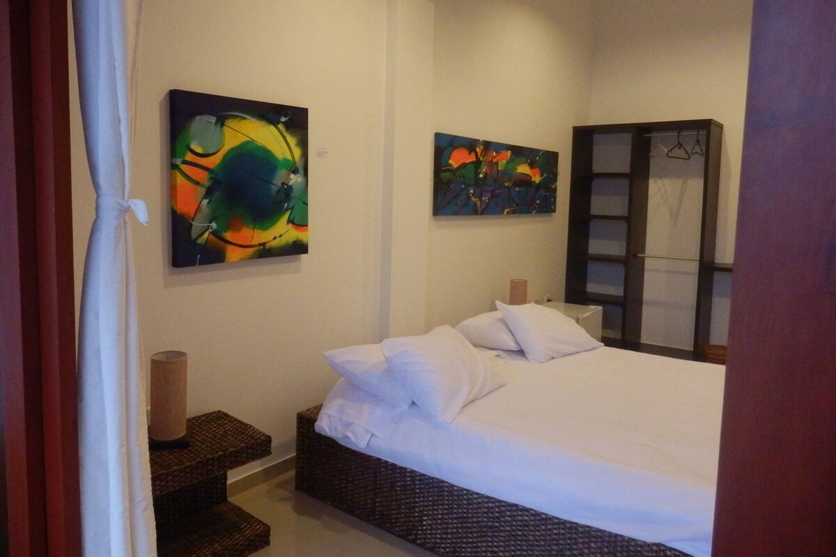 Un dormitorio con una cama, dos cuadros, un armario y una lámpara de noche.