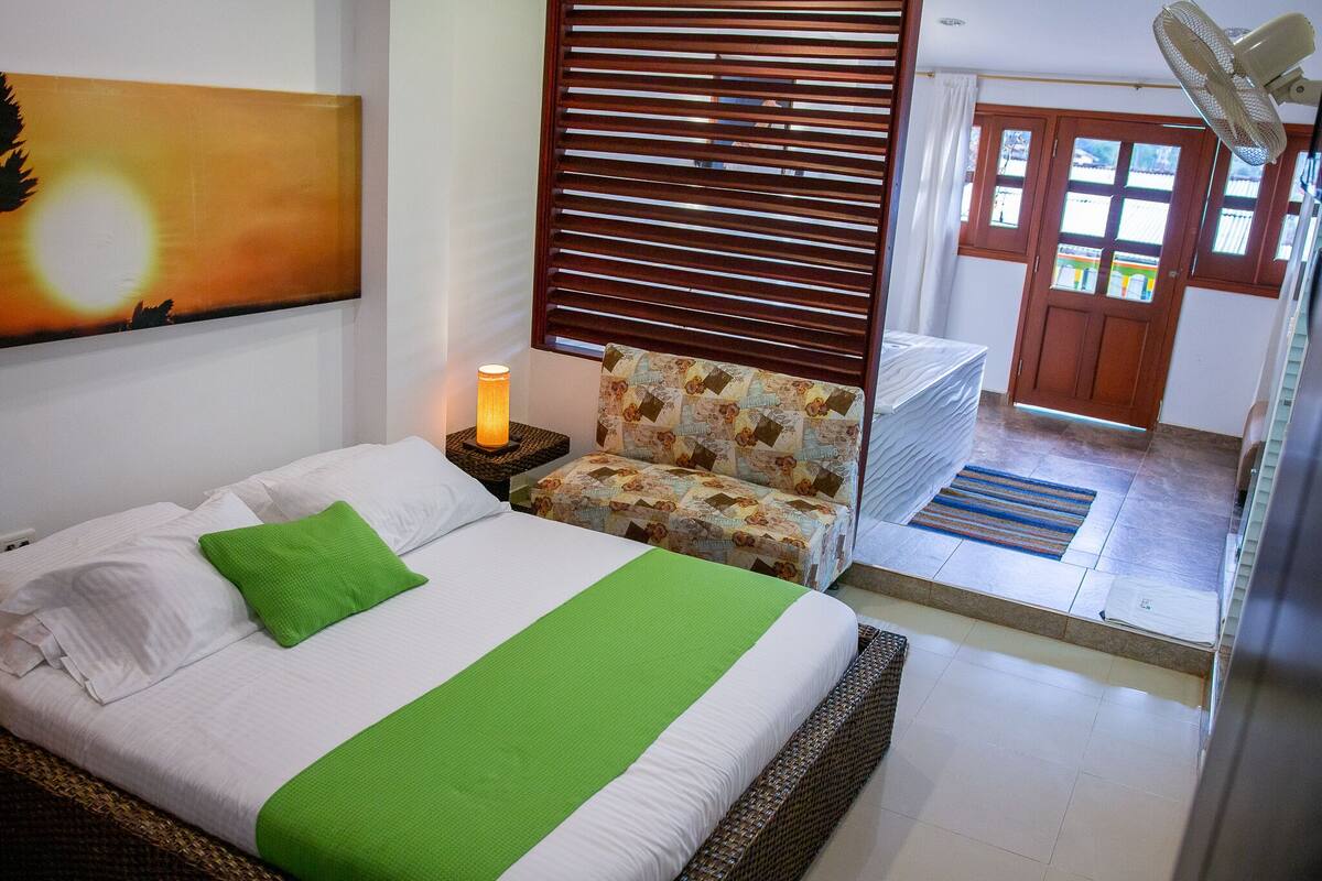 Una cama bien hecha con ropa de cama blanca y verde, un sillón estampado, un ventilador de pie y una puerta que da al exterior.
