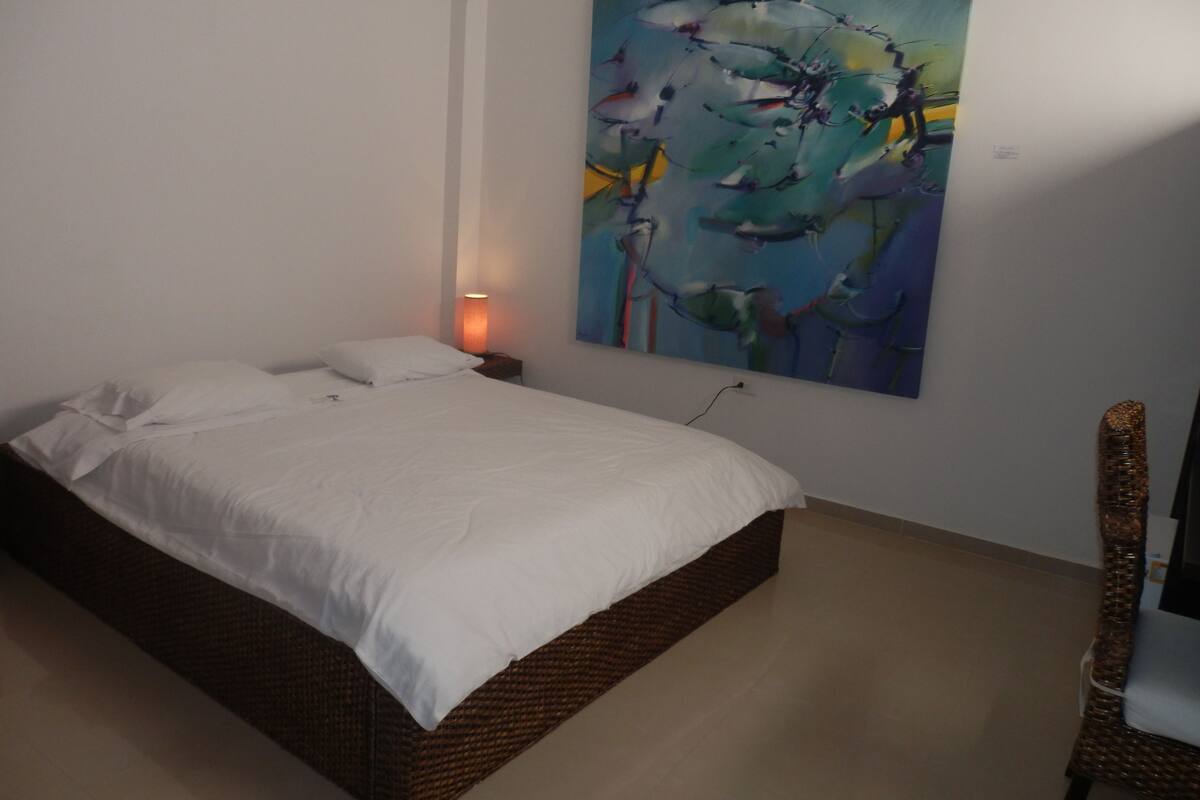 Un dormitorio con cama, mesita de noche, lámpara, silla y un cuadro abstracto grande en la pared.