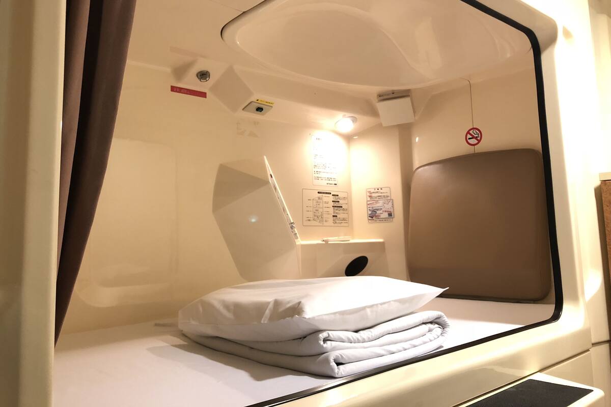 A small, private sleeping area with a bed, a pillow, a headboard, a window, and a wall-mounted light fixture.