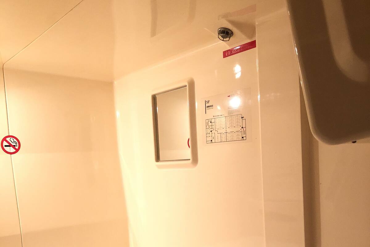 A hotel room with a white bed, a small window, and a no-smoking sign.