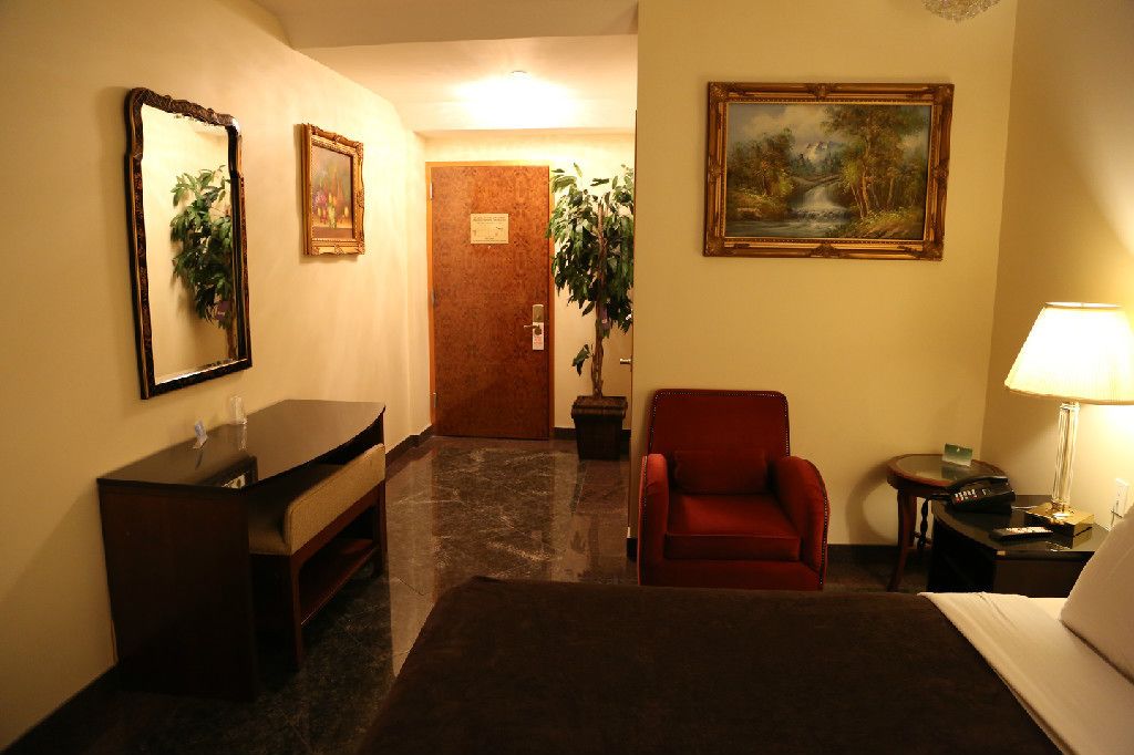 Una habitación de hotel con una cama, un escritorio, una silla, una lámpara y dos cuadros en las paredes.