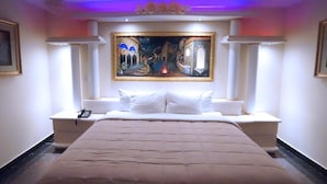 A hotel room with a large bed, white headboard, and a painting above the bed. - Paris Suites Hotel (Corona)
