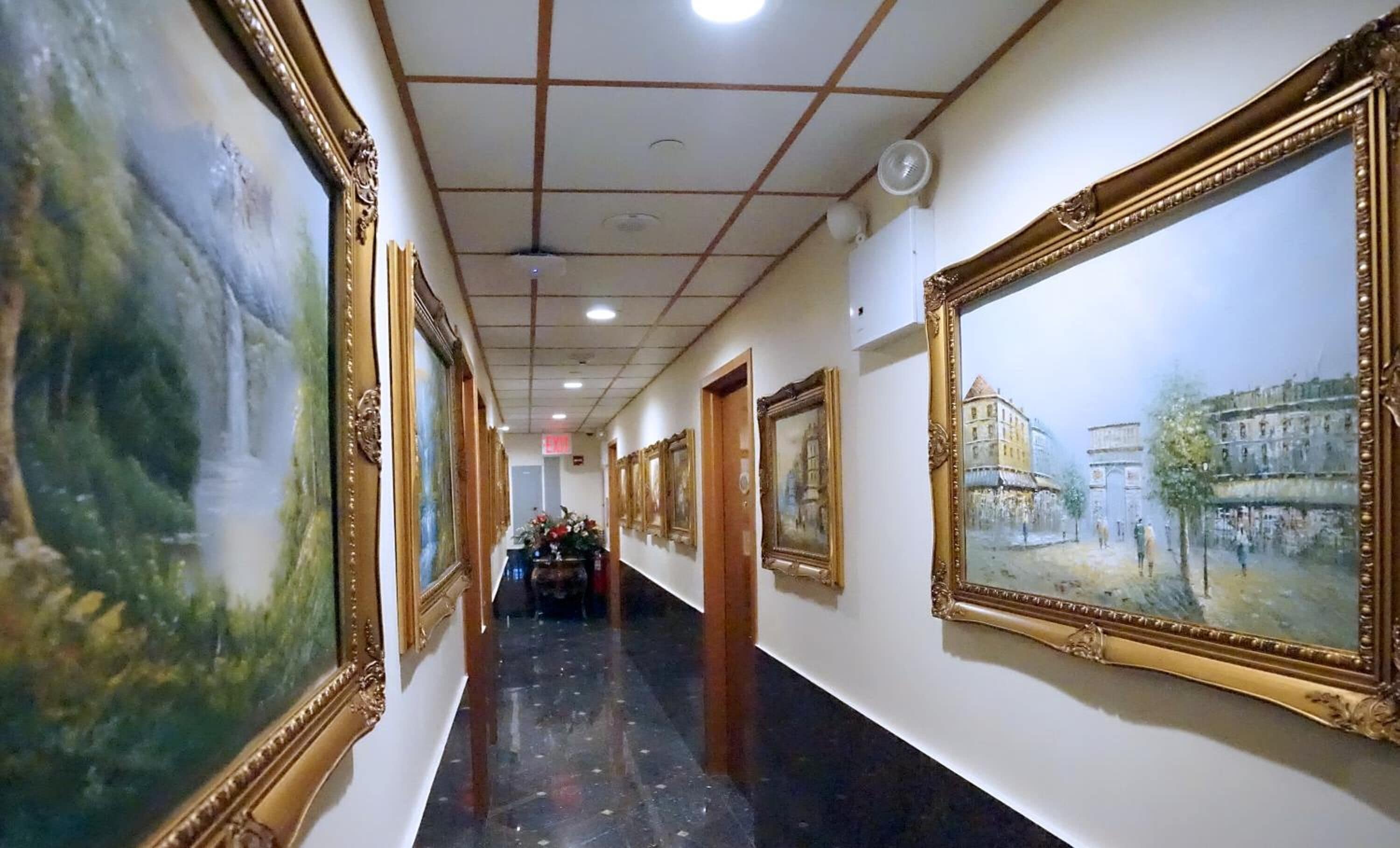 A corridor with framed paintings on the walls, a tiled floor, and a ceiling with recessed lighting.