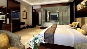 Une chambre d’hôtel dotée d’un grand lit, d’un coin salon avec un fauteuil, d’une télévision et d’une salle de bain fermée par des parois de verre.