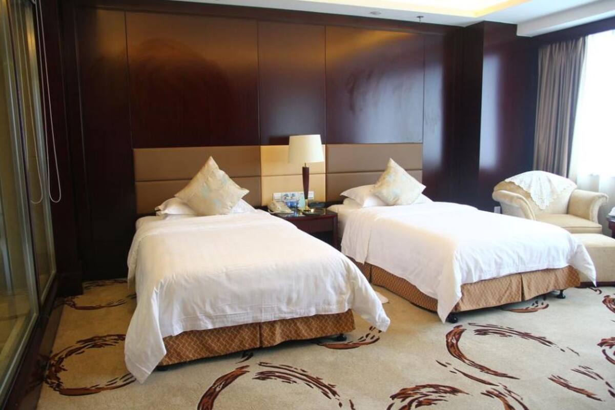 A hotel room with two beds, a bedside table with a lamp, a chair, and a large window.
